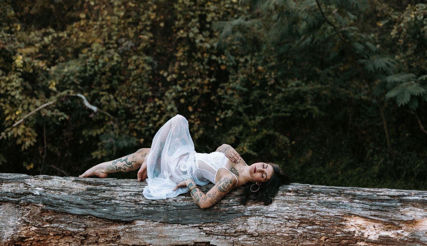 A tattooed woman in a white dress reclines gracefully on a mossy log surrounded by lush green forest.