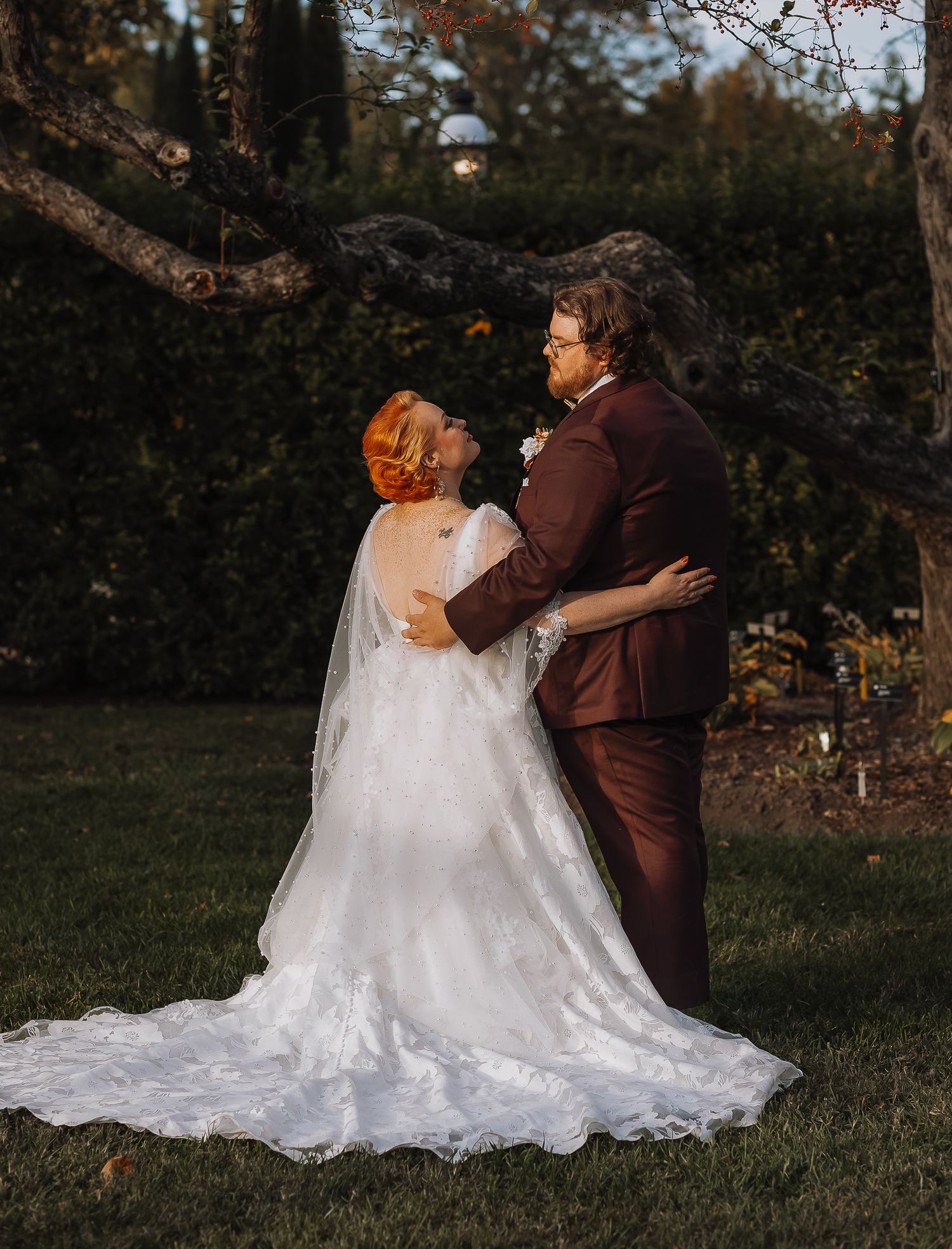 Couple in burgundy suit and white wedding dress embrace in garden at dusk.
