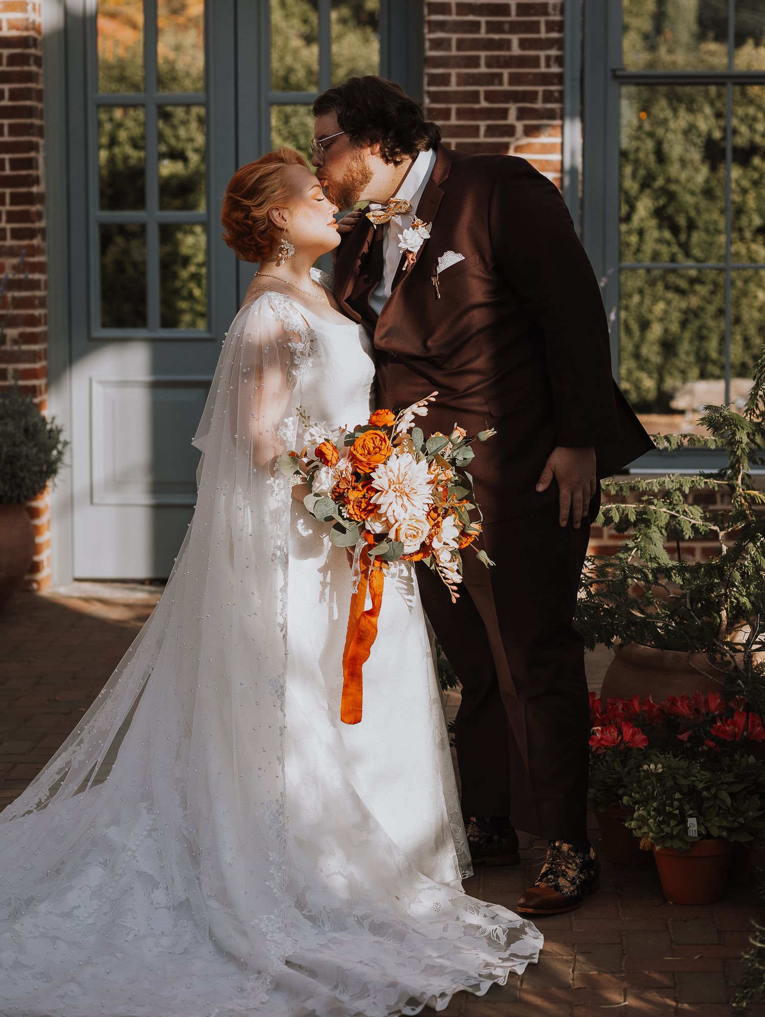 Bride in white dress holding orange bouquet shares romantic moment with groom in brown suit.
