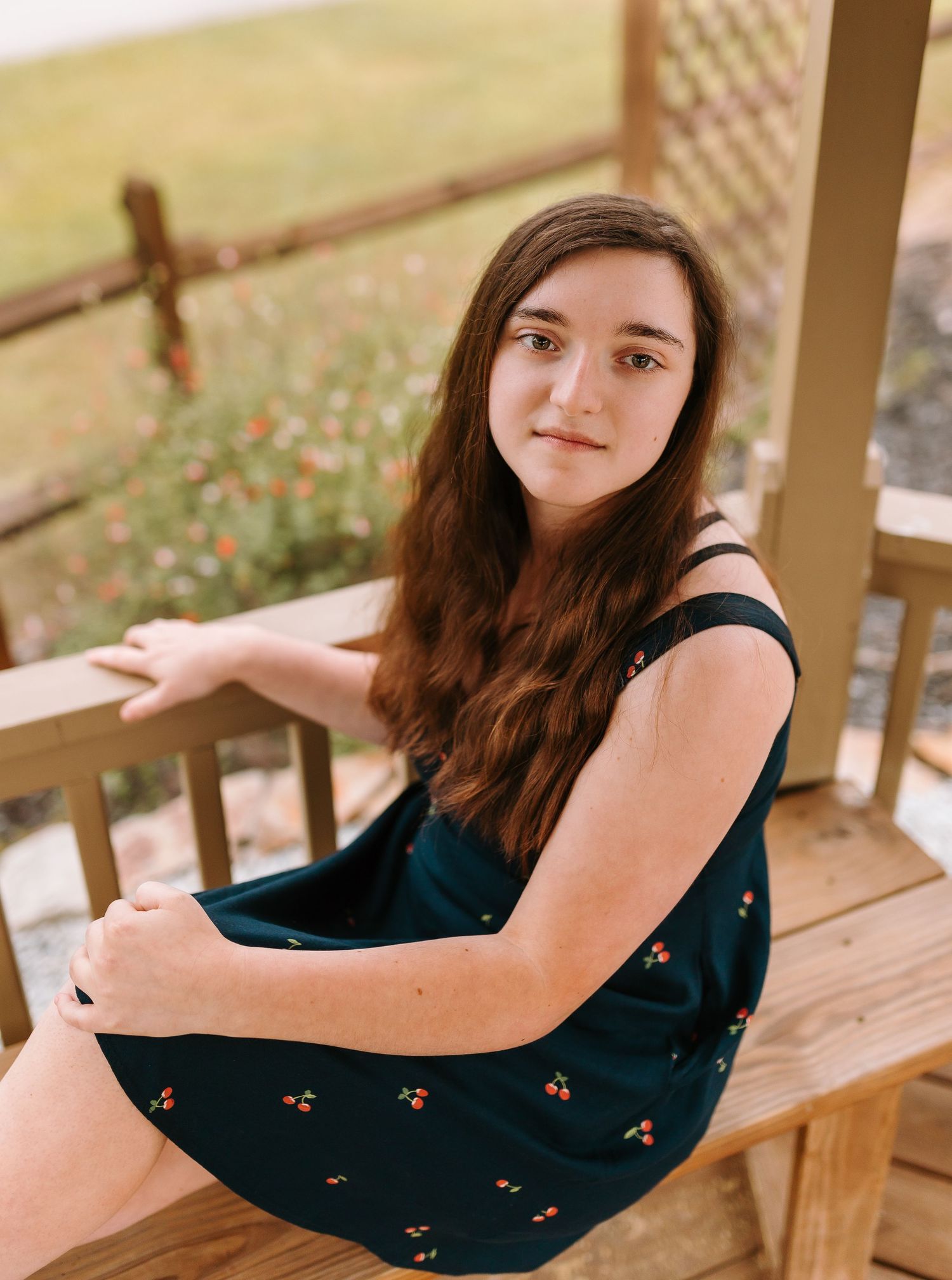 Young woman with long brown hair in a teal floral dress sitting on a wooden porch, smiling outdoors.