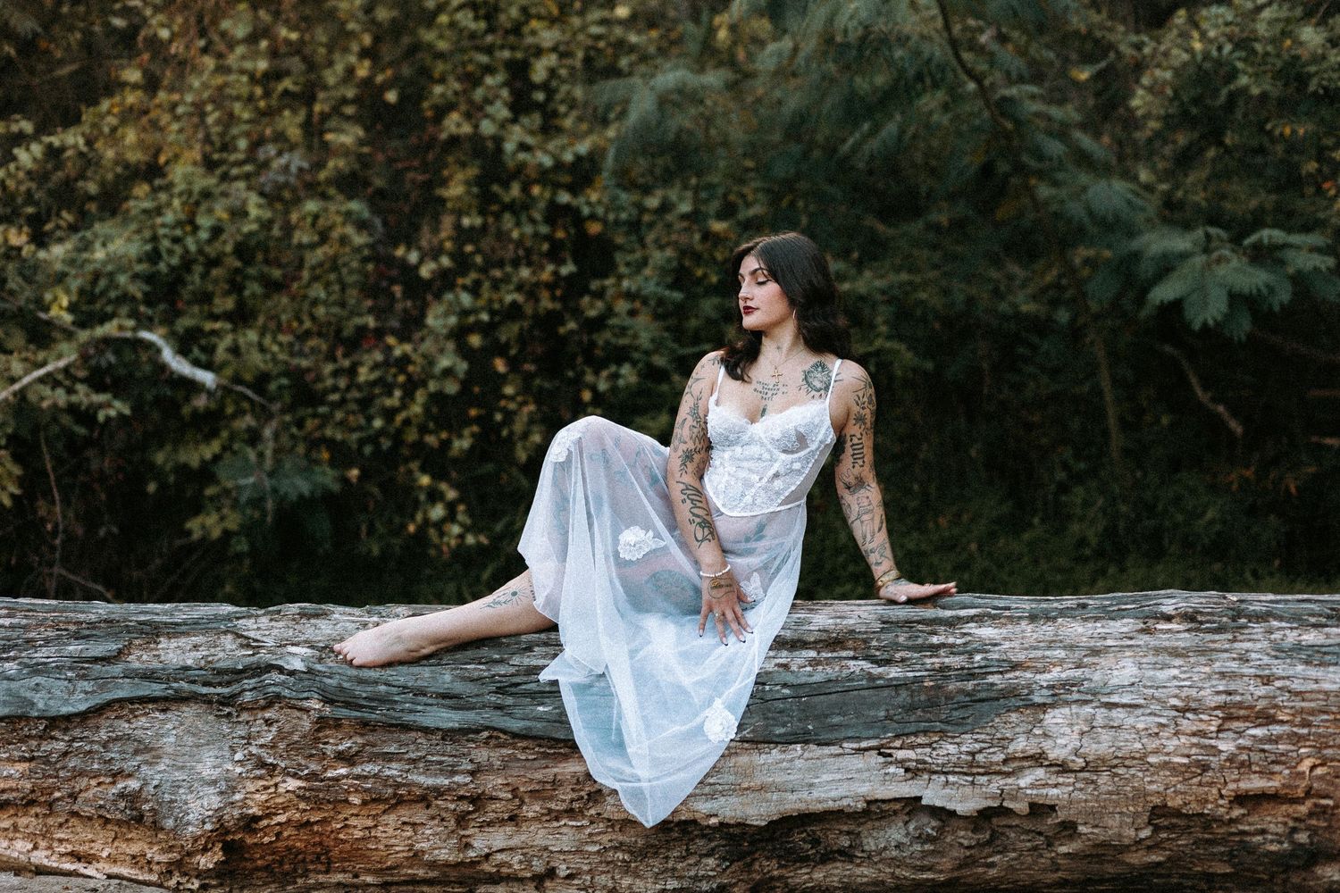 Tattooed woman in white lace dress sitting gracefully on a large log surrounded by lush green forest trees.