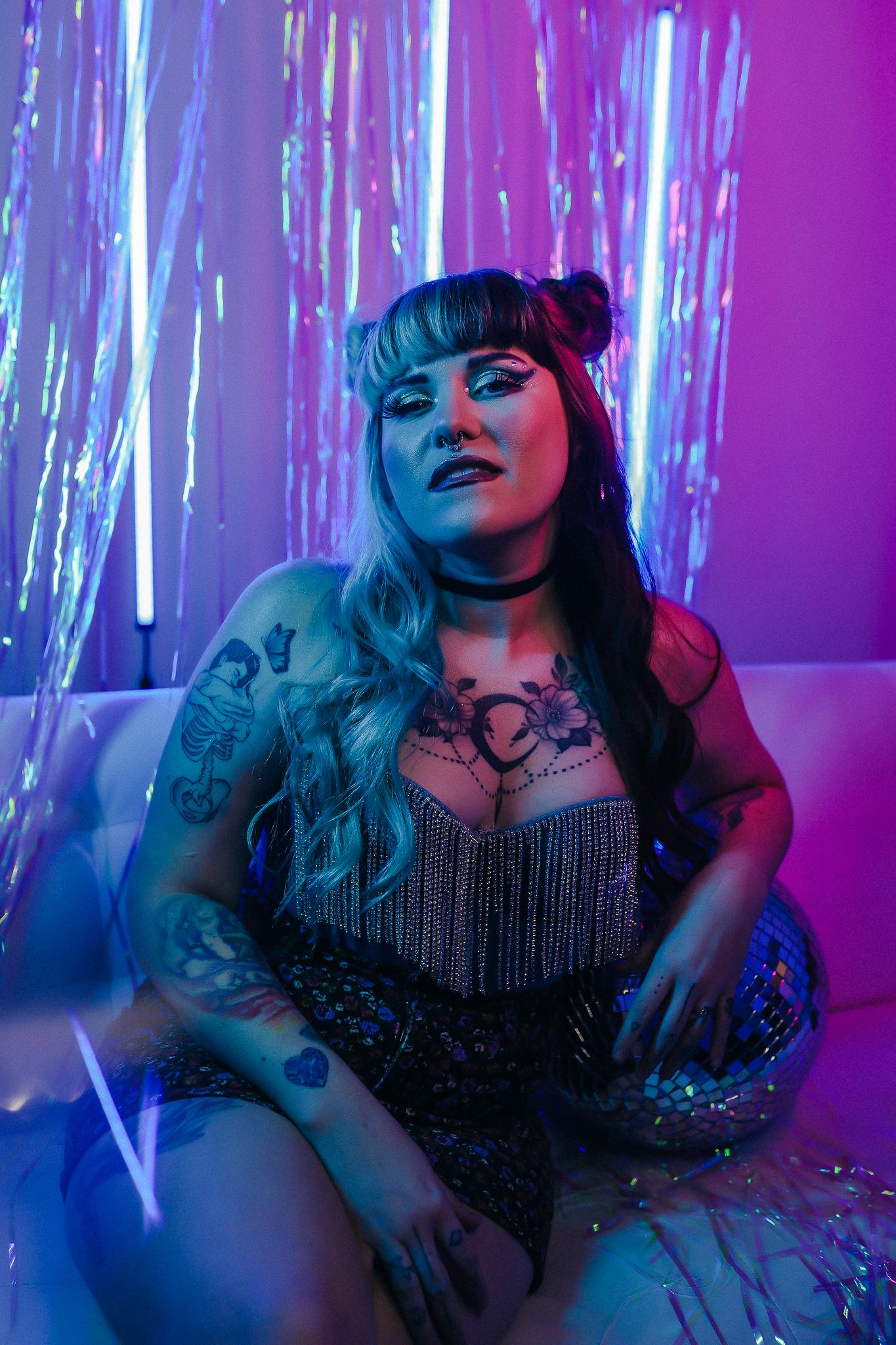 Tattooed woman in sparkly fringe dress poses on white couch under neon lights with holographic tinsel backdrop.