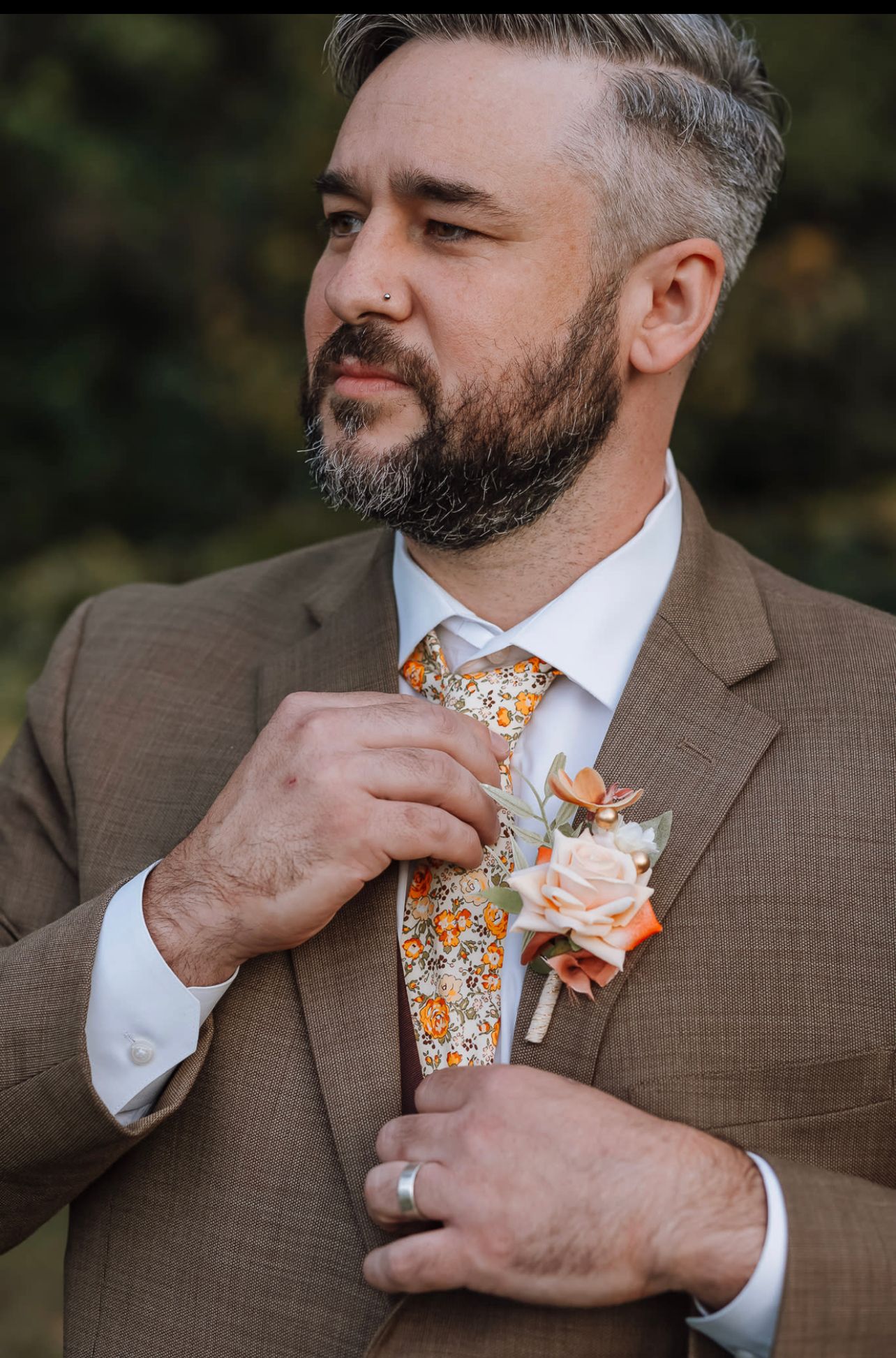 Groom adjusts floral tie while wearing brown suit with boutonniere.