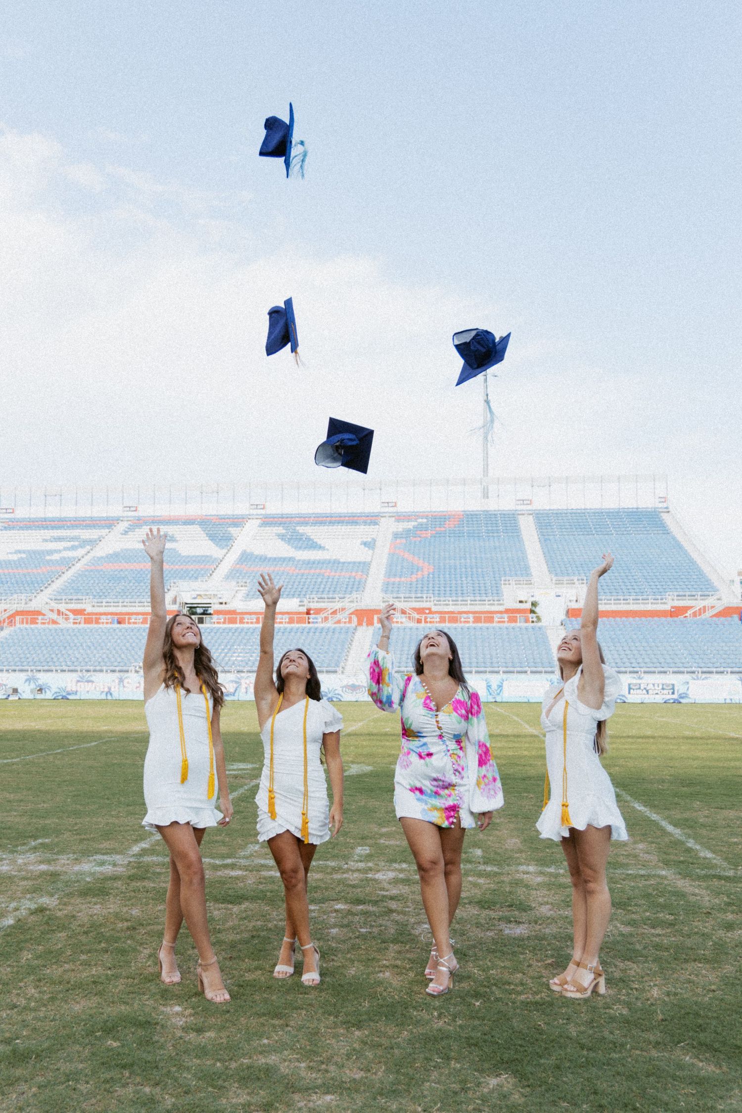 Graduates in white dresses tossing blue caps in the air on a football field.