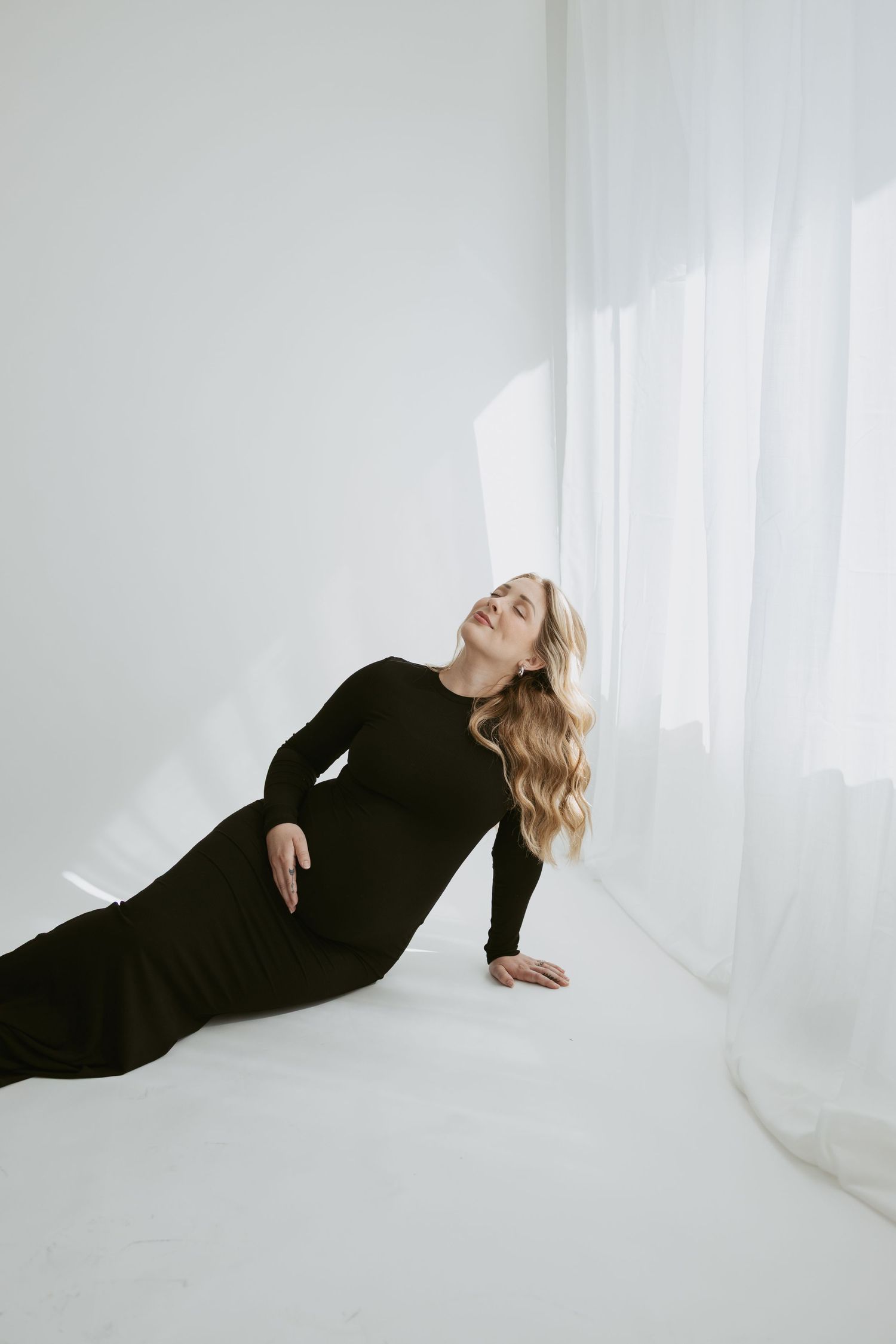 Artistic portrait of a woman in black dress reclining against white wall with dramatic lighting.