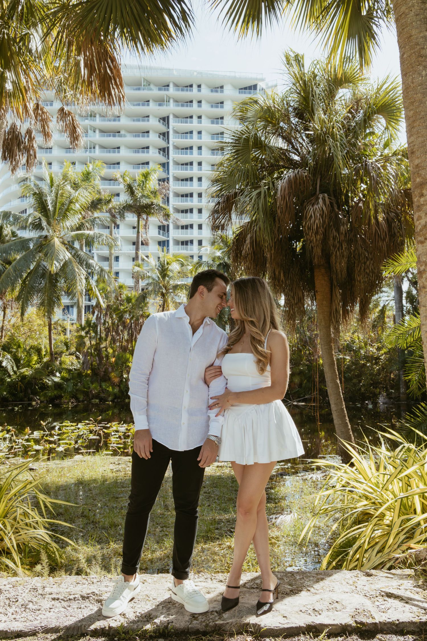 Couple in white outfits posing together near tropical resort with palm trees.