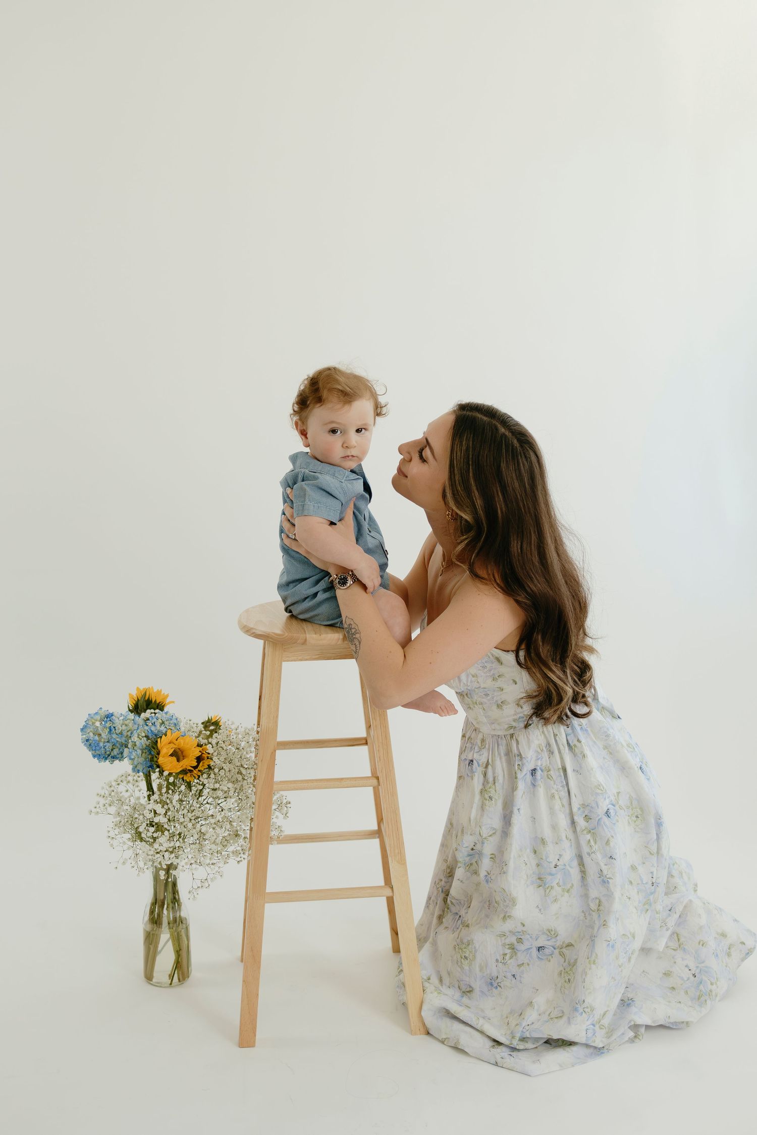 Parent and child share sweet moment by wooden stool with flower vase in white studio.
