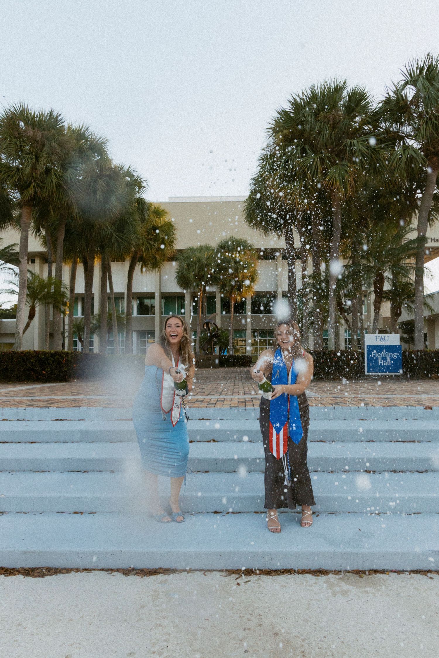 Two graduates celebrating with confetti on concrete steps surrounded by palm trees.