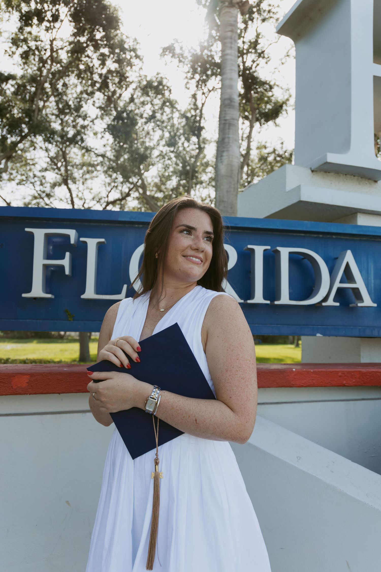 Graduate in white dress holds diploma while standing next to Florida university signage.