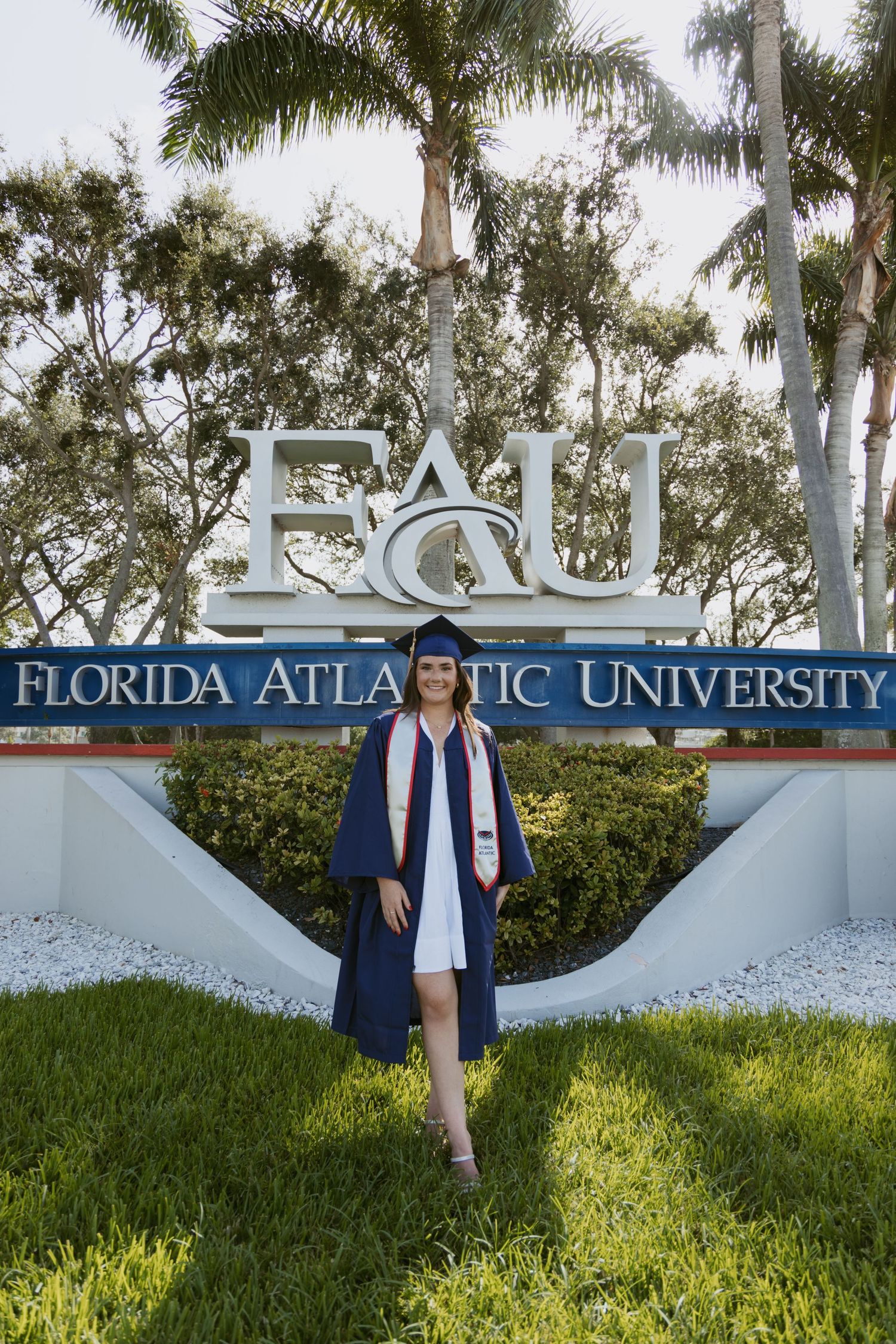 Graduate in cap and gown poses by FAU Florida Atlantic University entrance sign surrounded by palm trees.