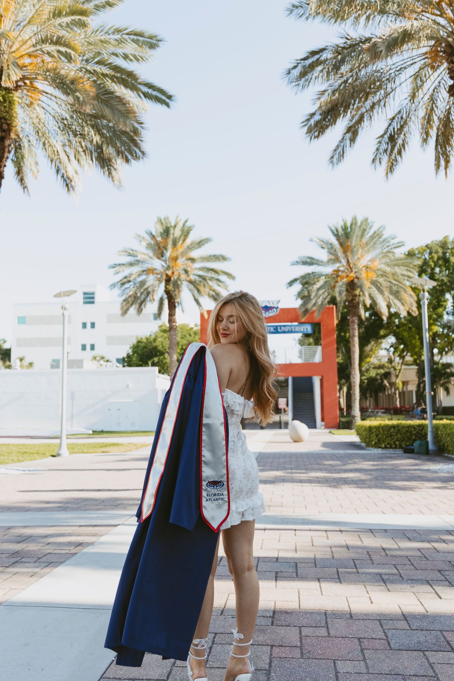 Graduate in white dress posing with graduation stoles under palm trees.