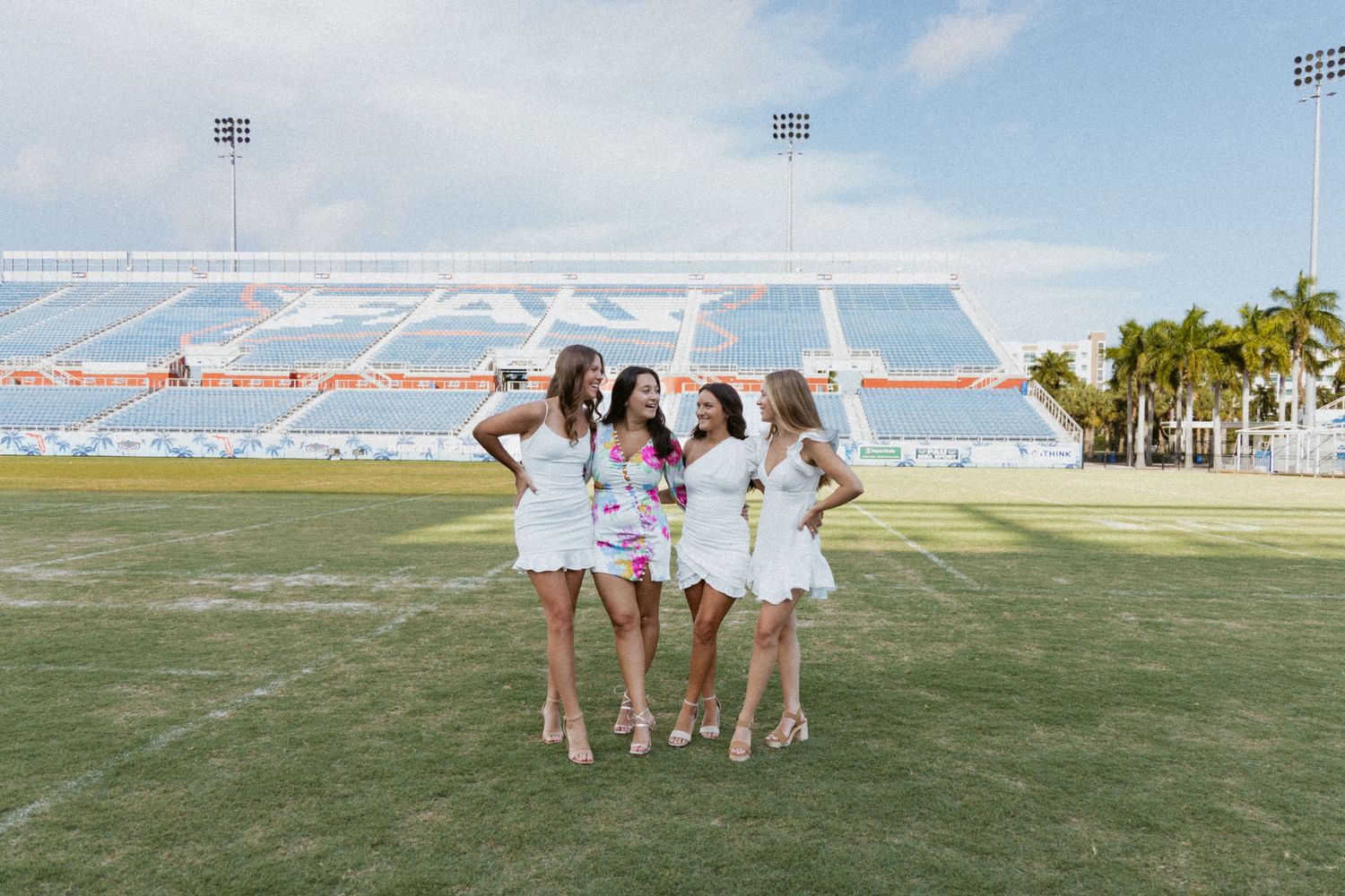 Friends in white dresses celebrating graduation on a football field with stadium seating.