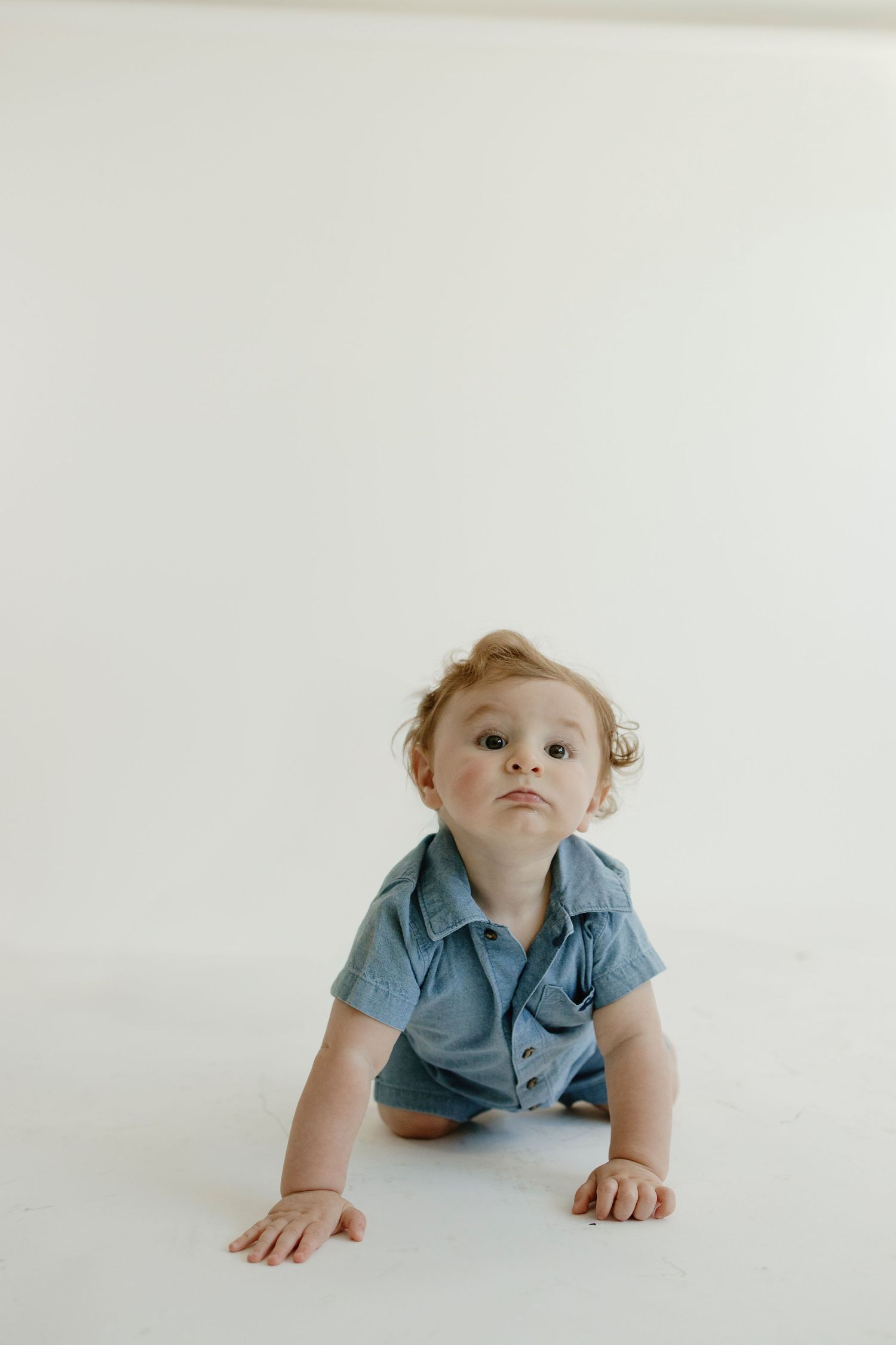 Baby in blue shirt crawling on white studio floor looking up at camera.