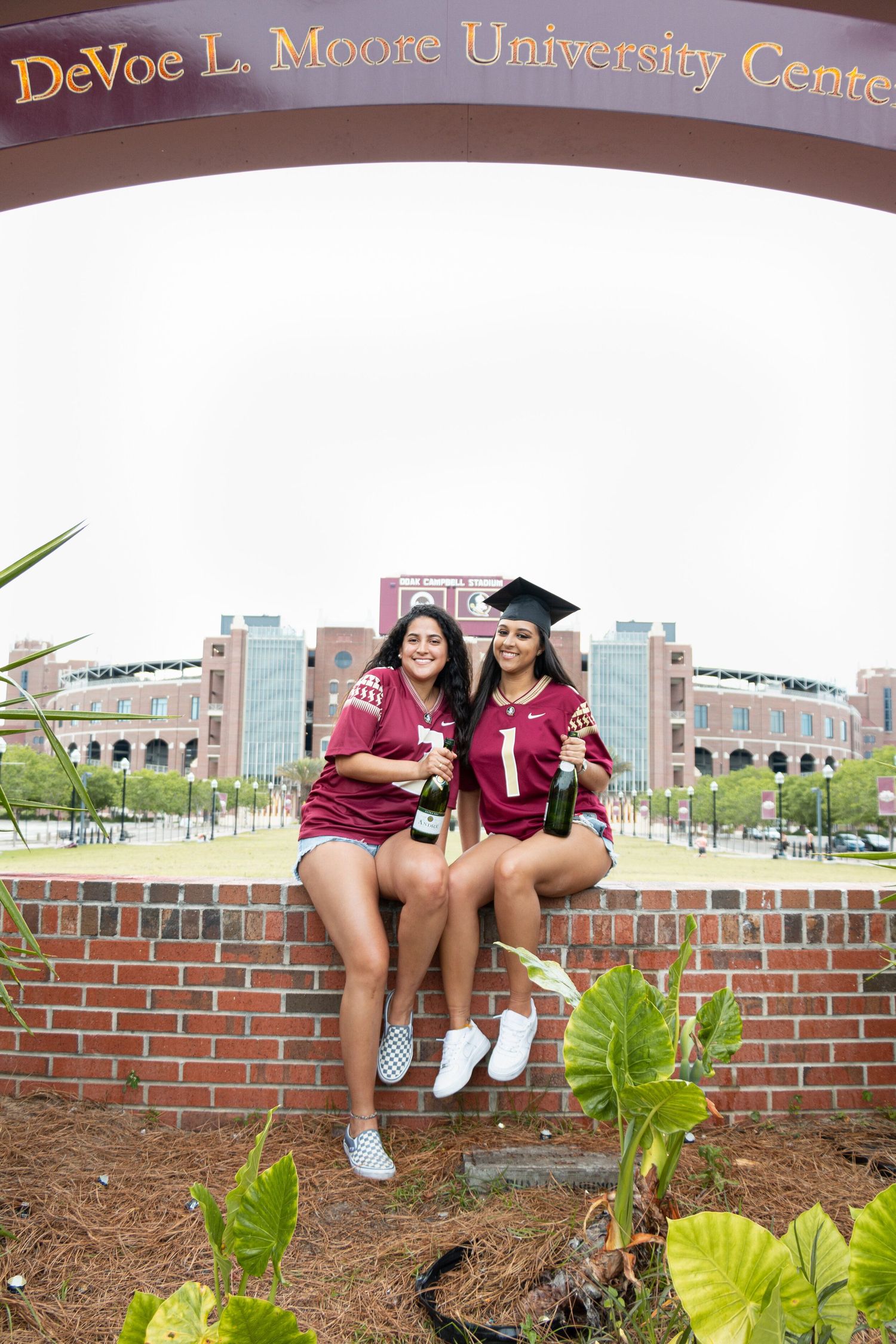 Two graduates in maroon and white pose at DeVoe L. Moore University Center entrance.