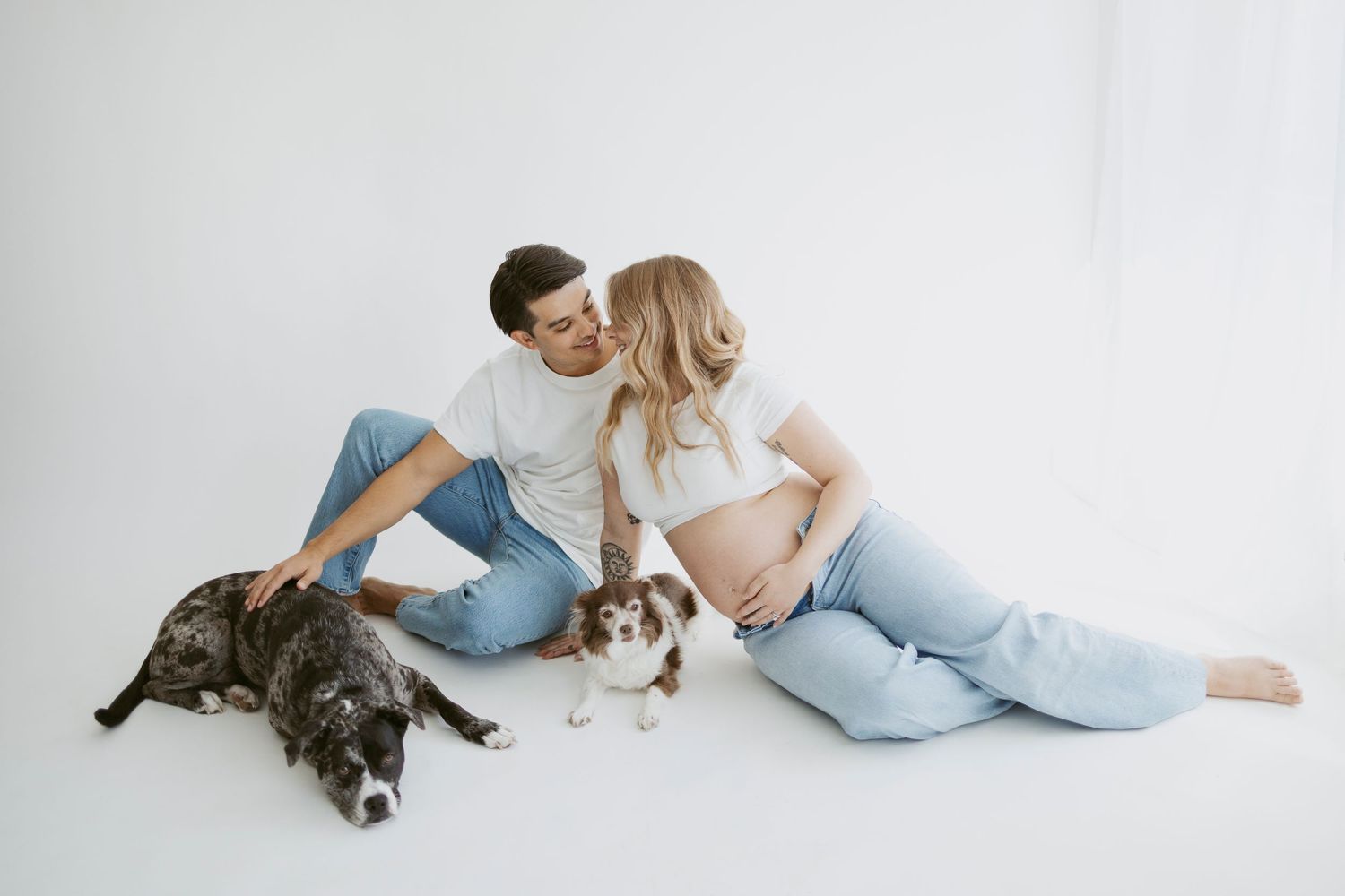 Expecting couple sits with their pets on white floor during maternity photoshoot.