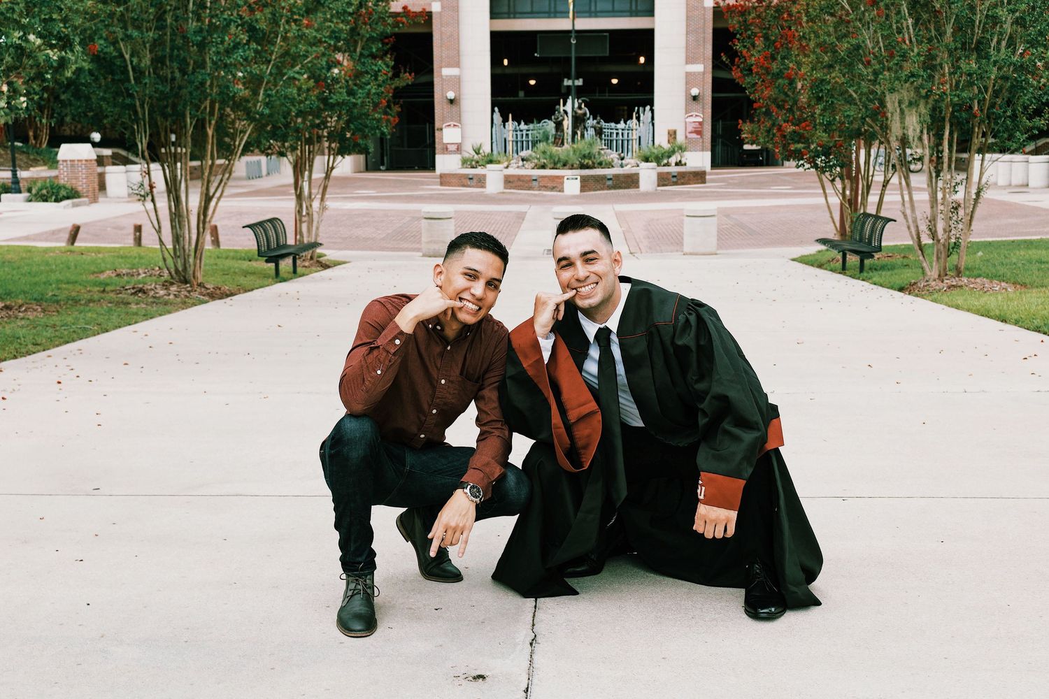 Two graduates in black robes pose together on campus walkway.