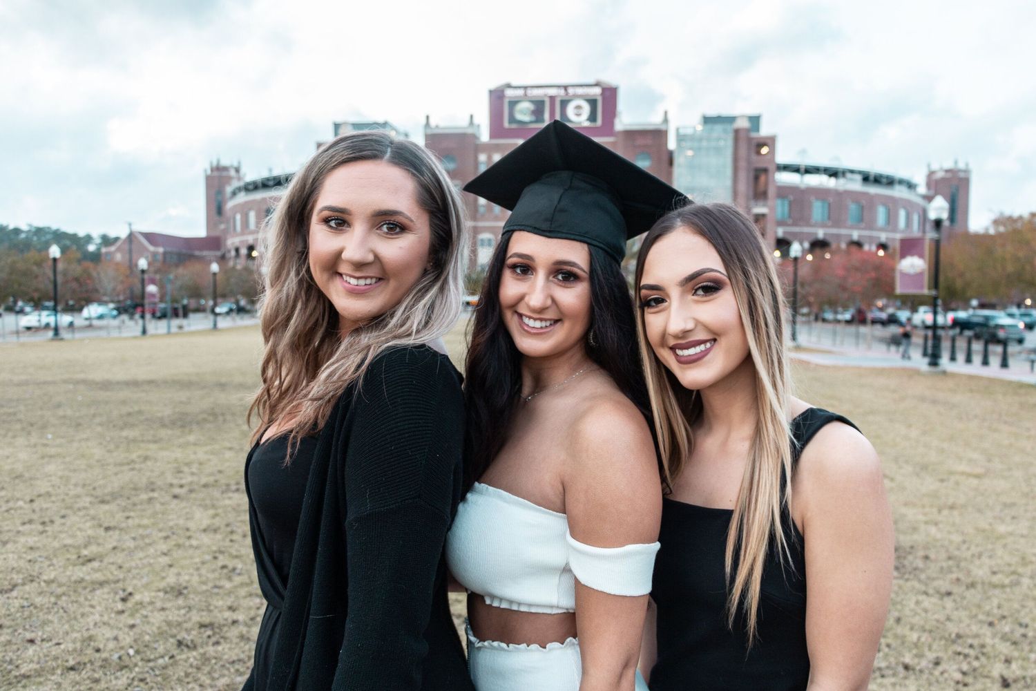 Three graduates posing together on campus grounds with buildings in the background.