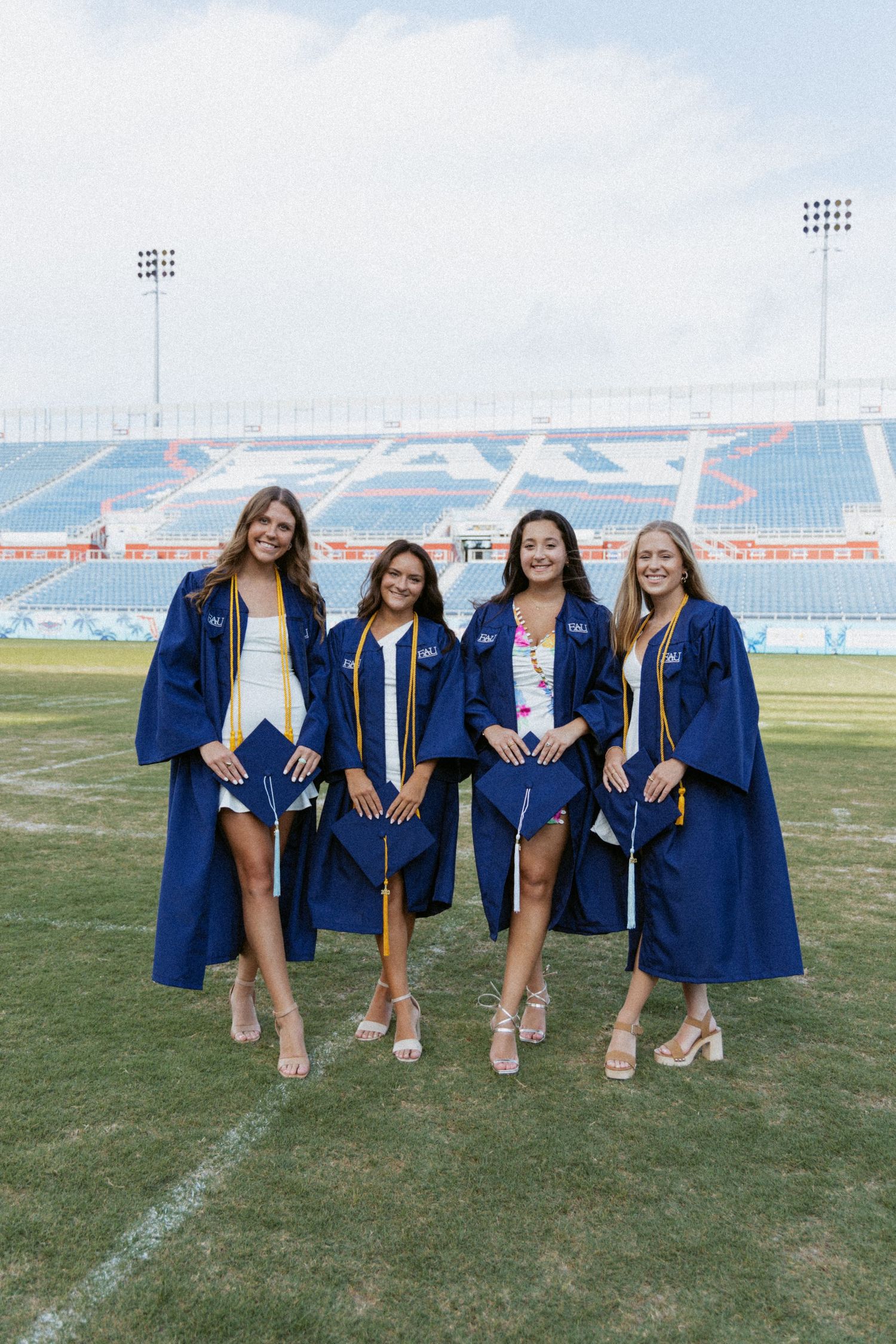 Graduates wearing blue commencement robes pose together on a football field with stadium seating in the background.