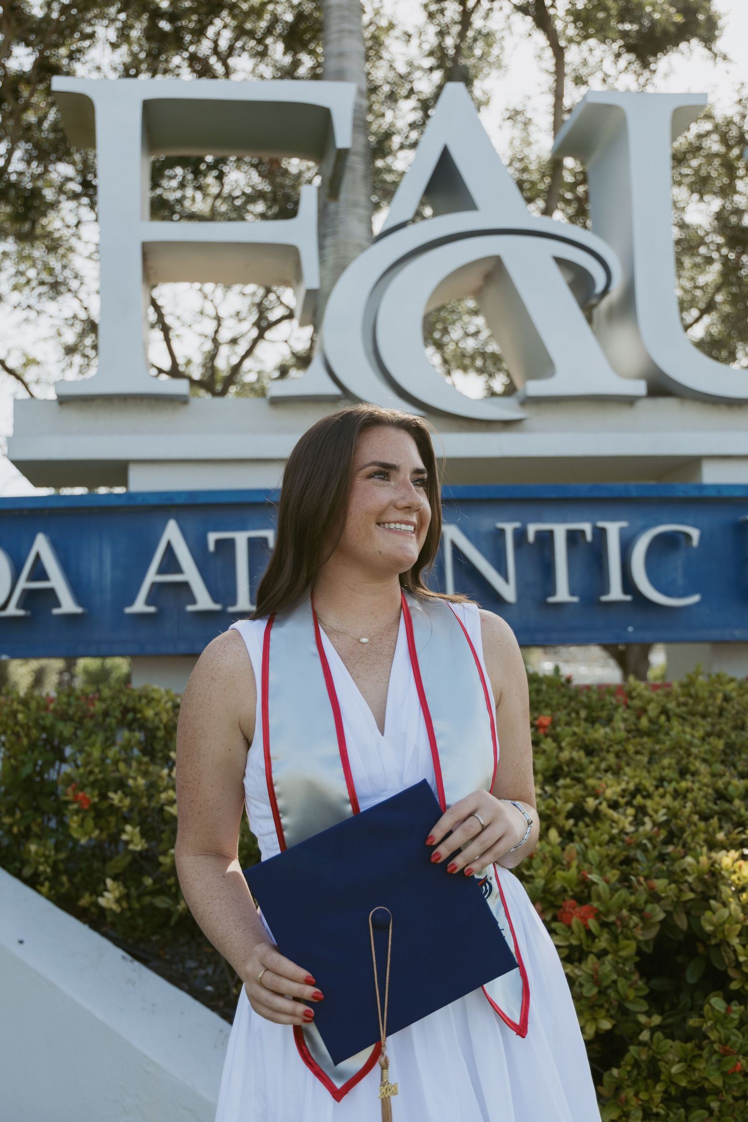 Graduate in a white dress holding diploma poses in front of Florida Atlantic University sign.