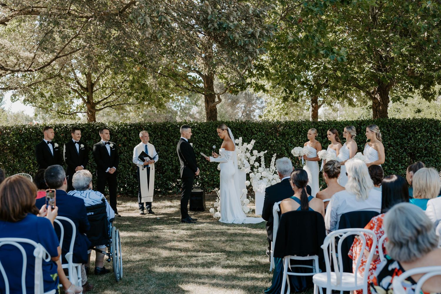 Outdoor garden wedding ceremony with guests seated in white chairs under shady trees during daytime.