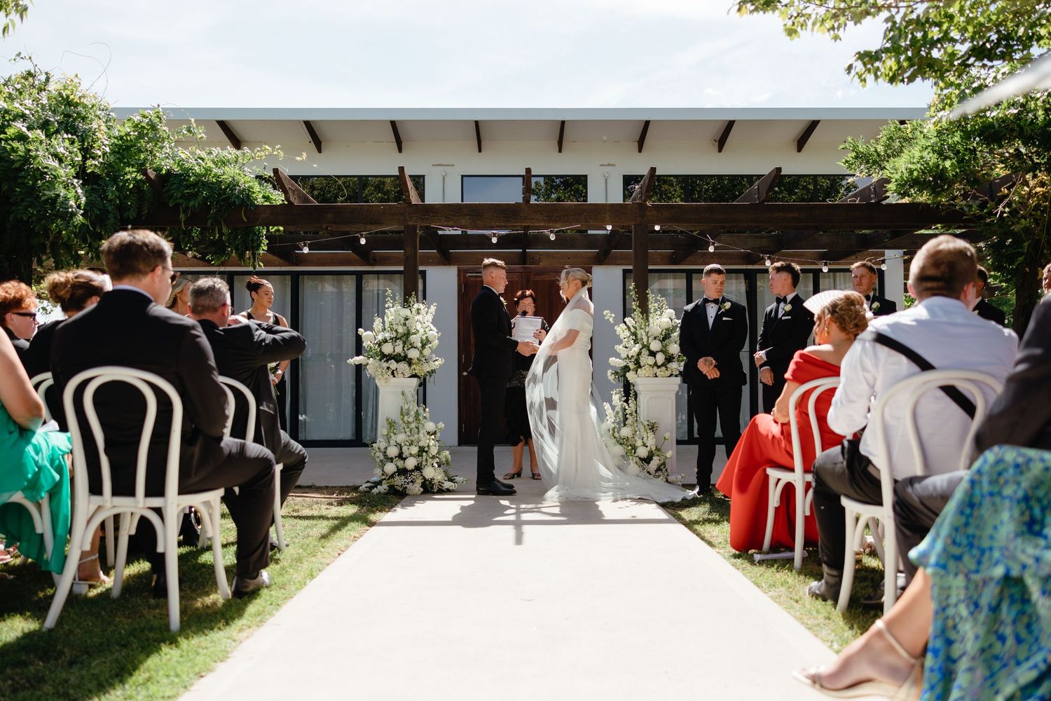 Wedding guests mingle and socialize under an elegant white pergola during an outdoor summer reception.