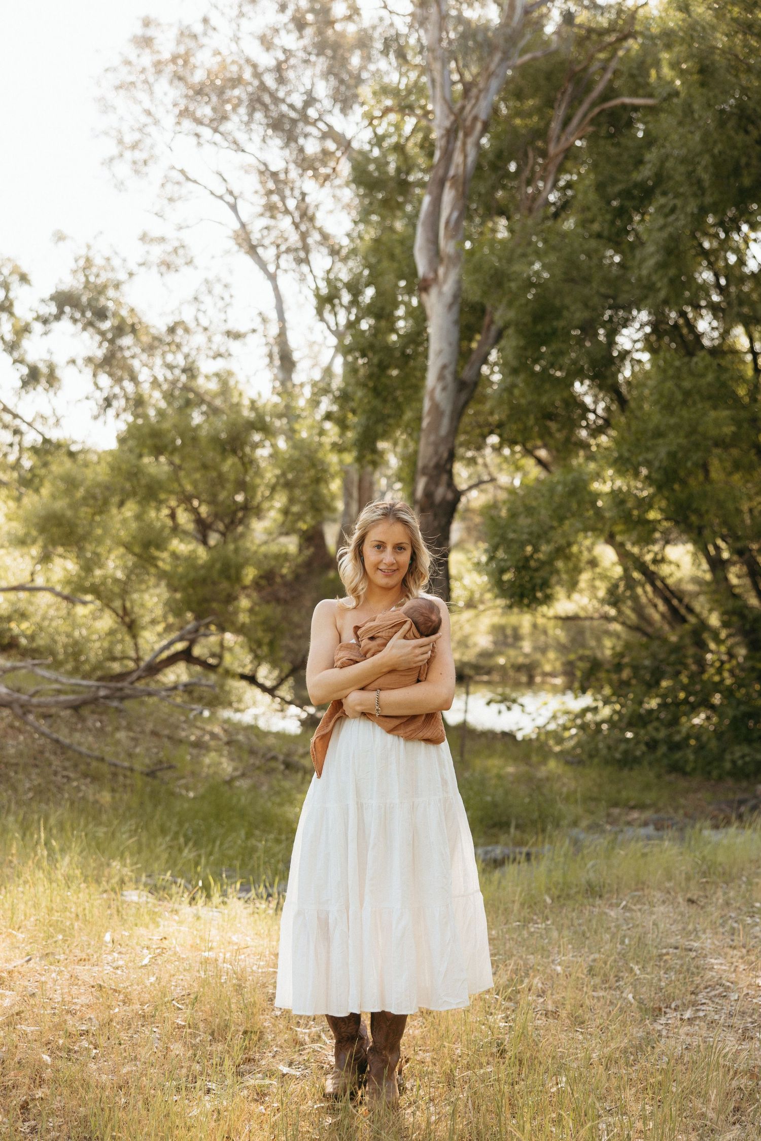 A person in a white dress holding a violin stands in a sunlit meadow with trees in the background.