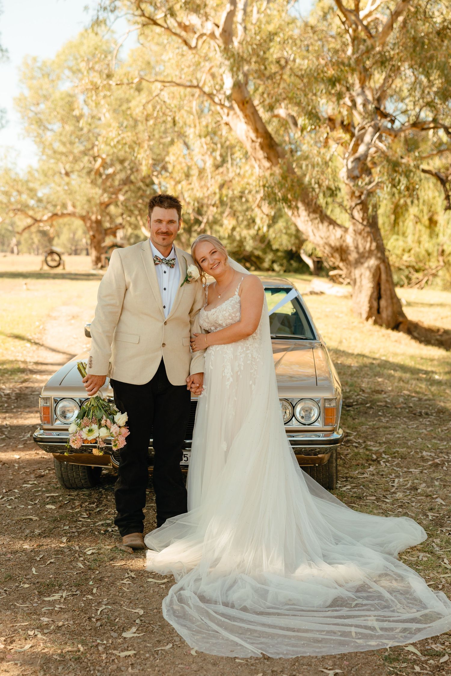 A newlywed couple poses in front of a vintage car under golden sunlit trees with a flowing white veil and bouquet.