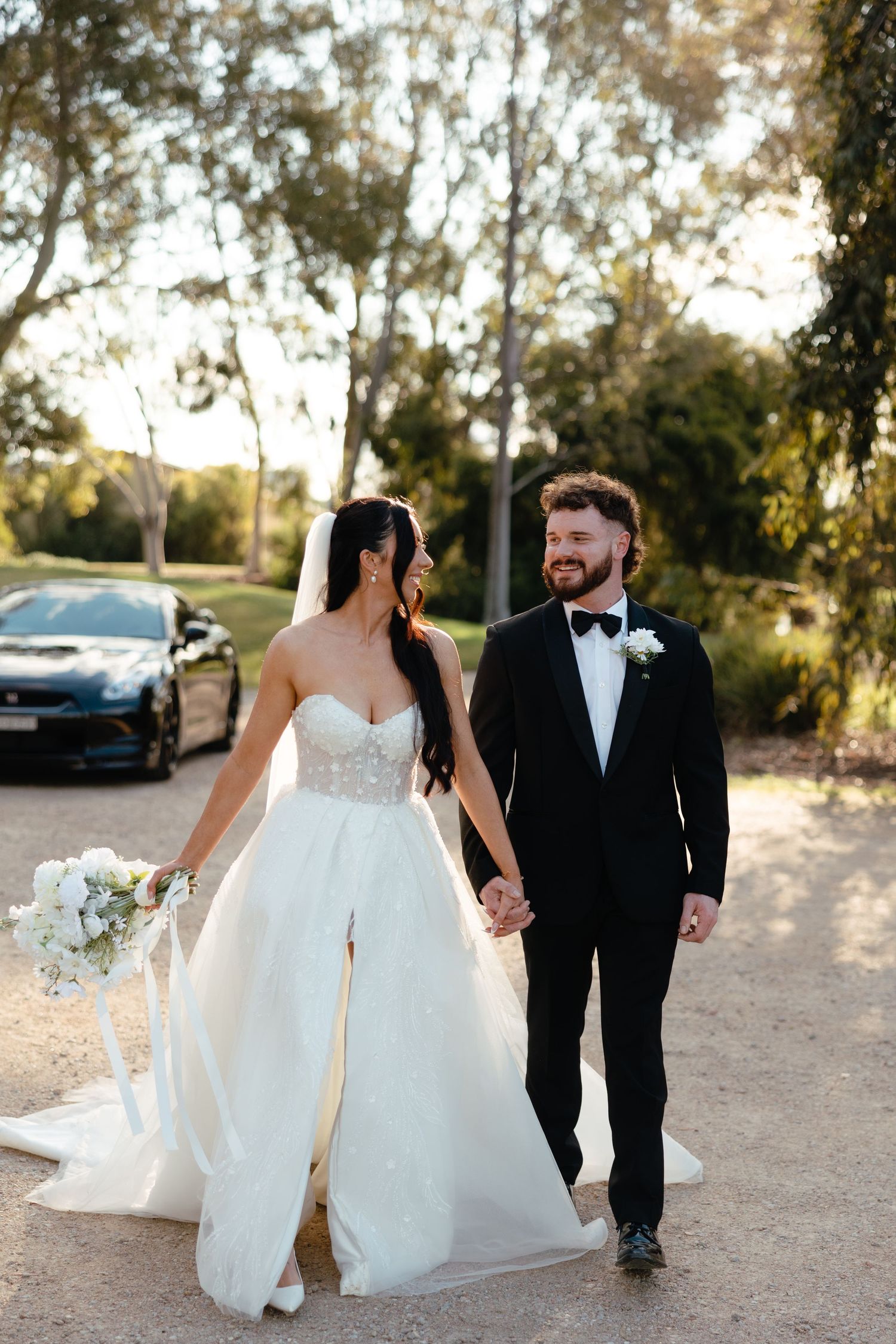 A romantic couple in formal wedding attire walk together on a dirt path during golden hour.