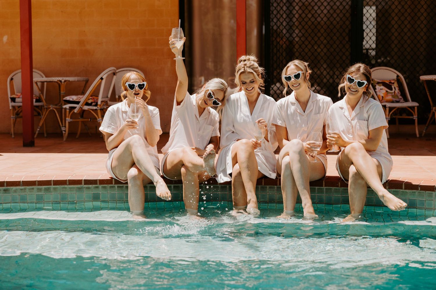 Five friends in matching white robes and sunglasses sit poolside with their feet dangling in turquoise water.