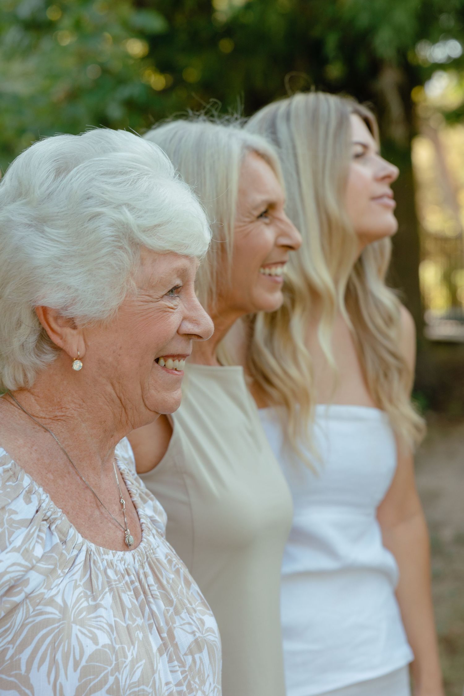 Three generations of women smiling together in sunlit outdoor setting.