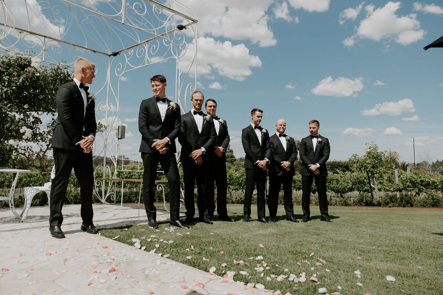 Groomsmen in black tuxedos stand on grass under white wedding arch against blue sky with puffy clouds.