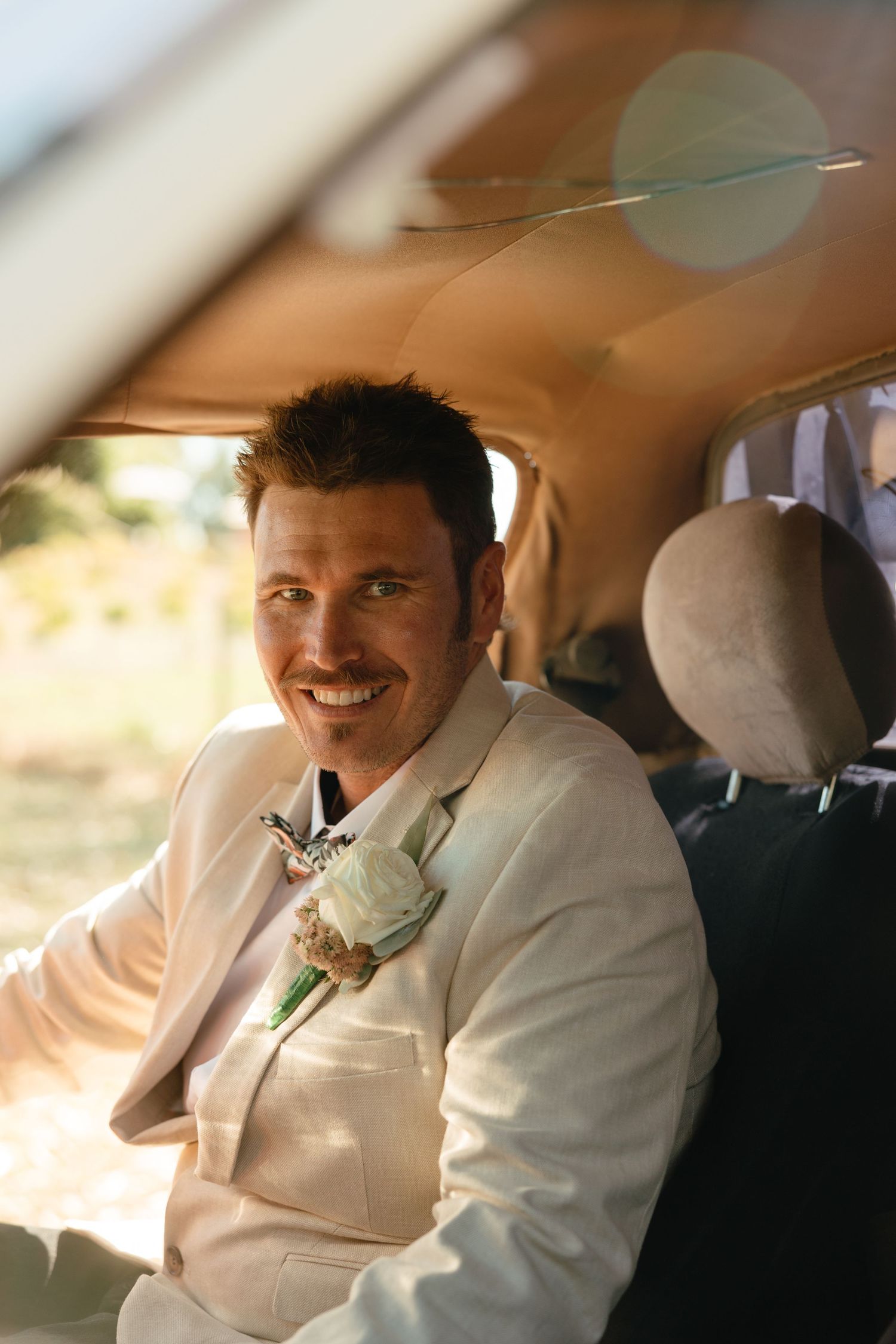 A person wearing a white suit smiles while sitting in the backseat of a car.