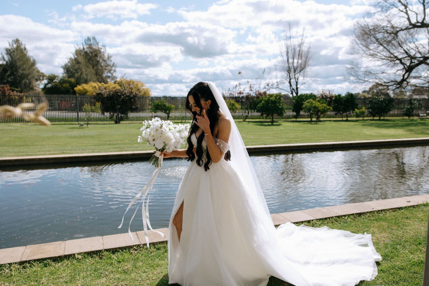 A scenic wedding couple poses by a serene lake surrounded by lush greenery and a cloudy sky at Lancemore Hotel Milawa