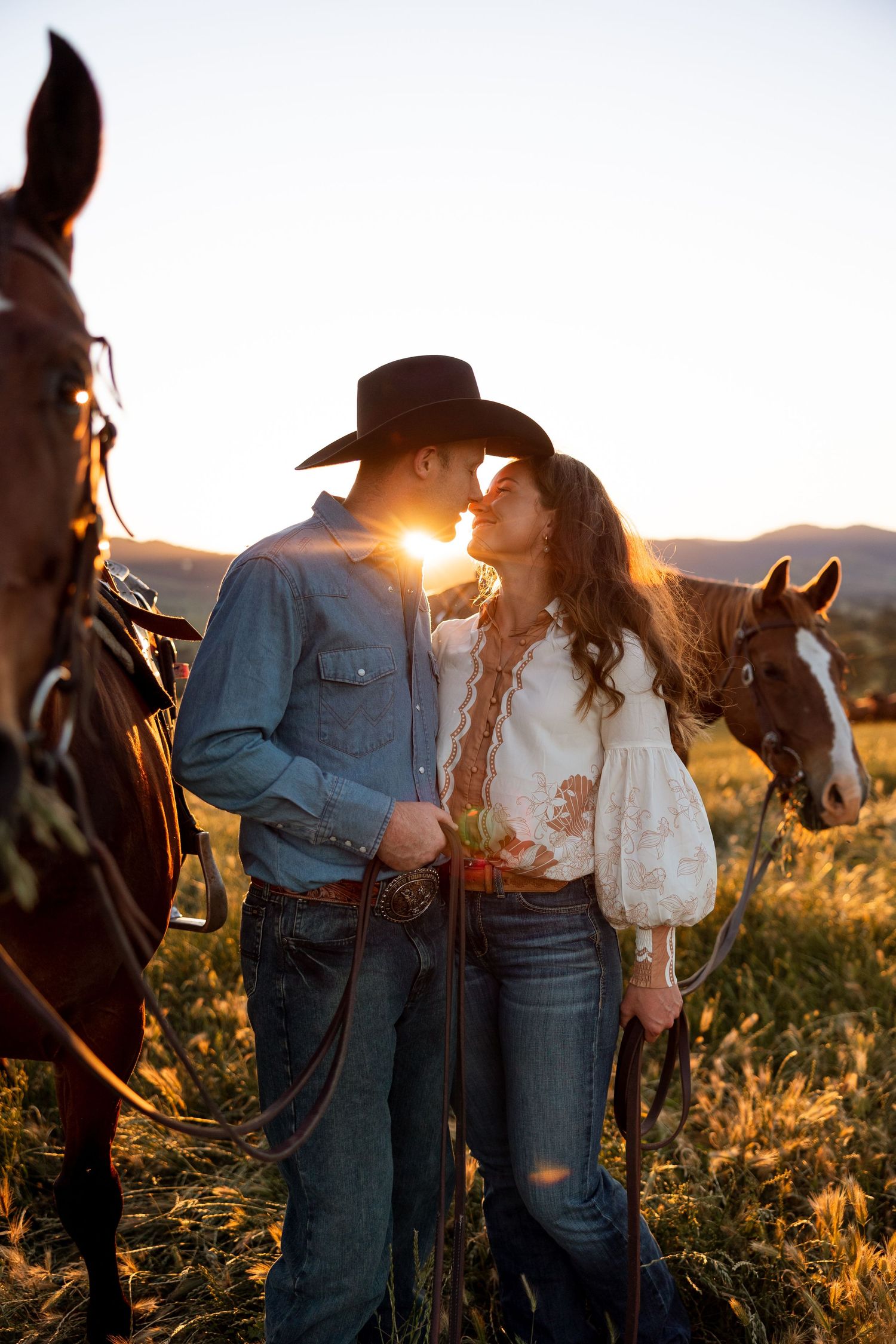 A romantic couple in western attire stands with their horse during golden hour sunset in a rustic outdoor setting.