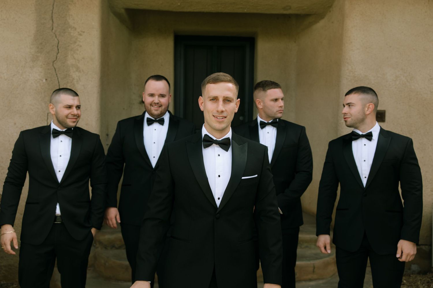 A group of groomsmen in black tuxedos and bowties stand together in a formal wedding party lineup.