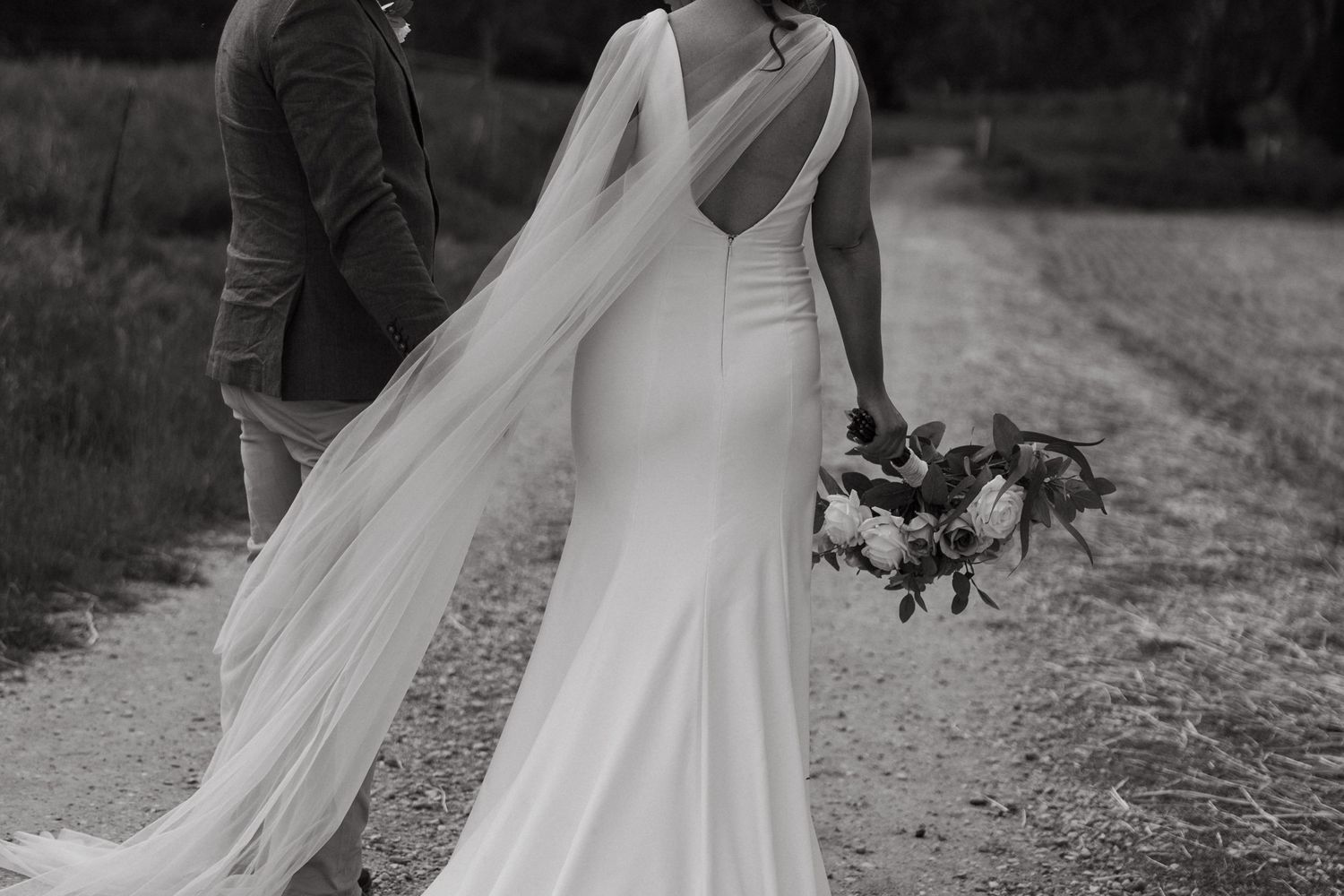 Elegant bride in fitted dress with flowing veil holds bouquet on country path.