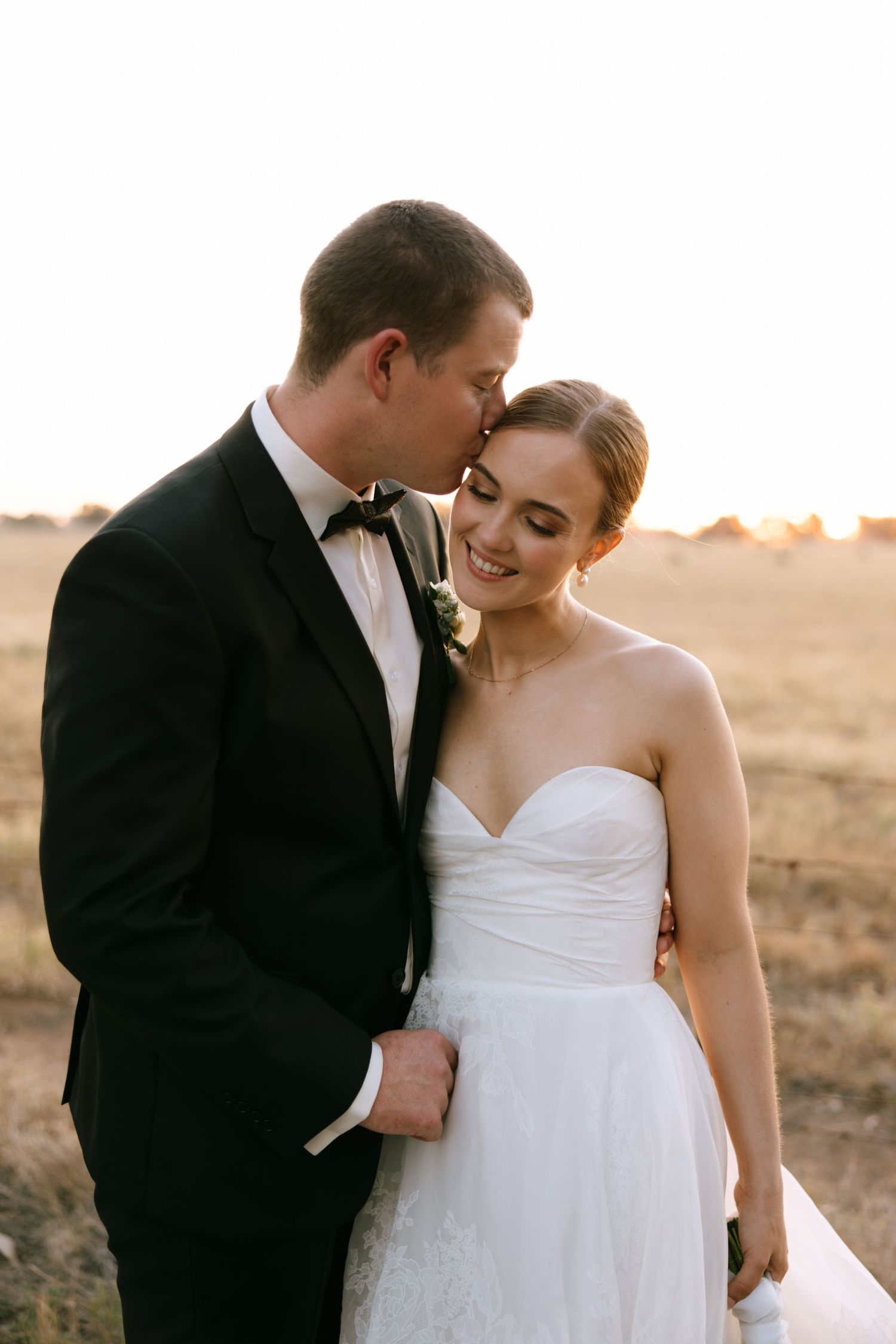 A tender moment between newlyweds at sunset, with the bride in white and groom in black tuxedo embracing in a field.