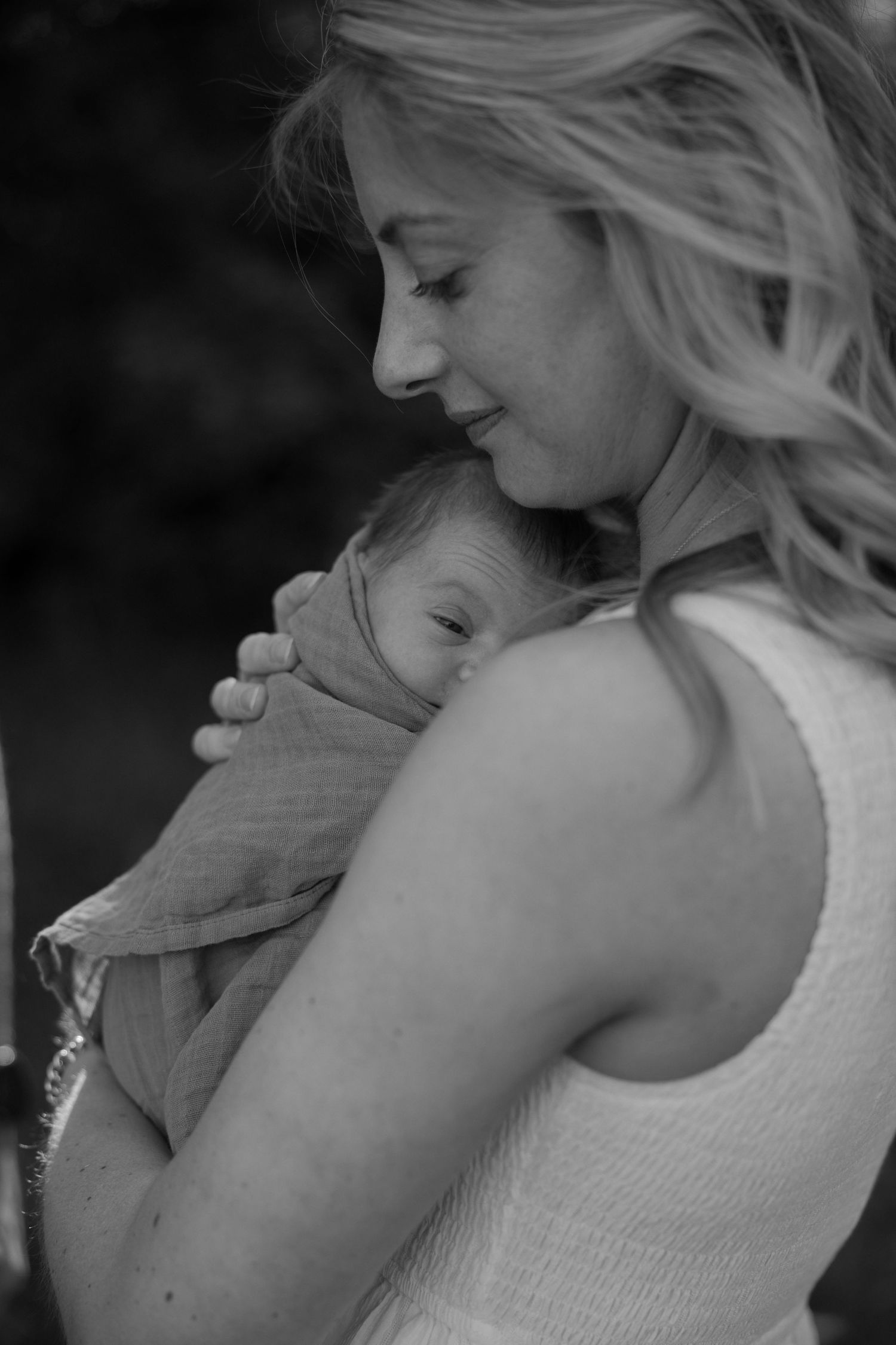 A tender black and white portrait of a mother in a sleeveless top cradling a sleeping infant against her chest.