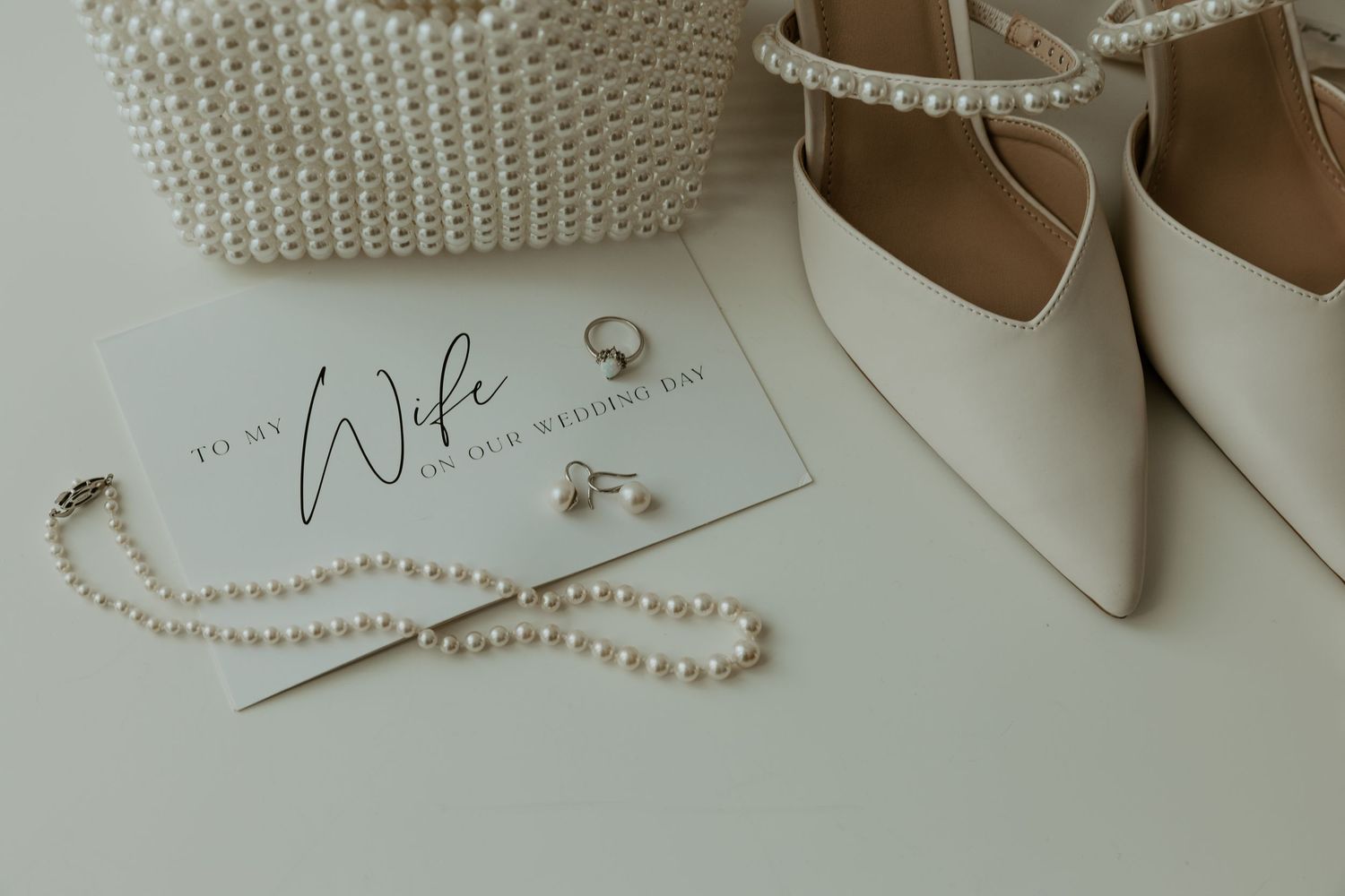 White pointed bridal shoes with pearl necklace and 'Wife' text card on a light colored surface.