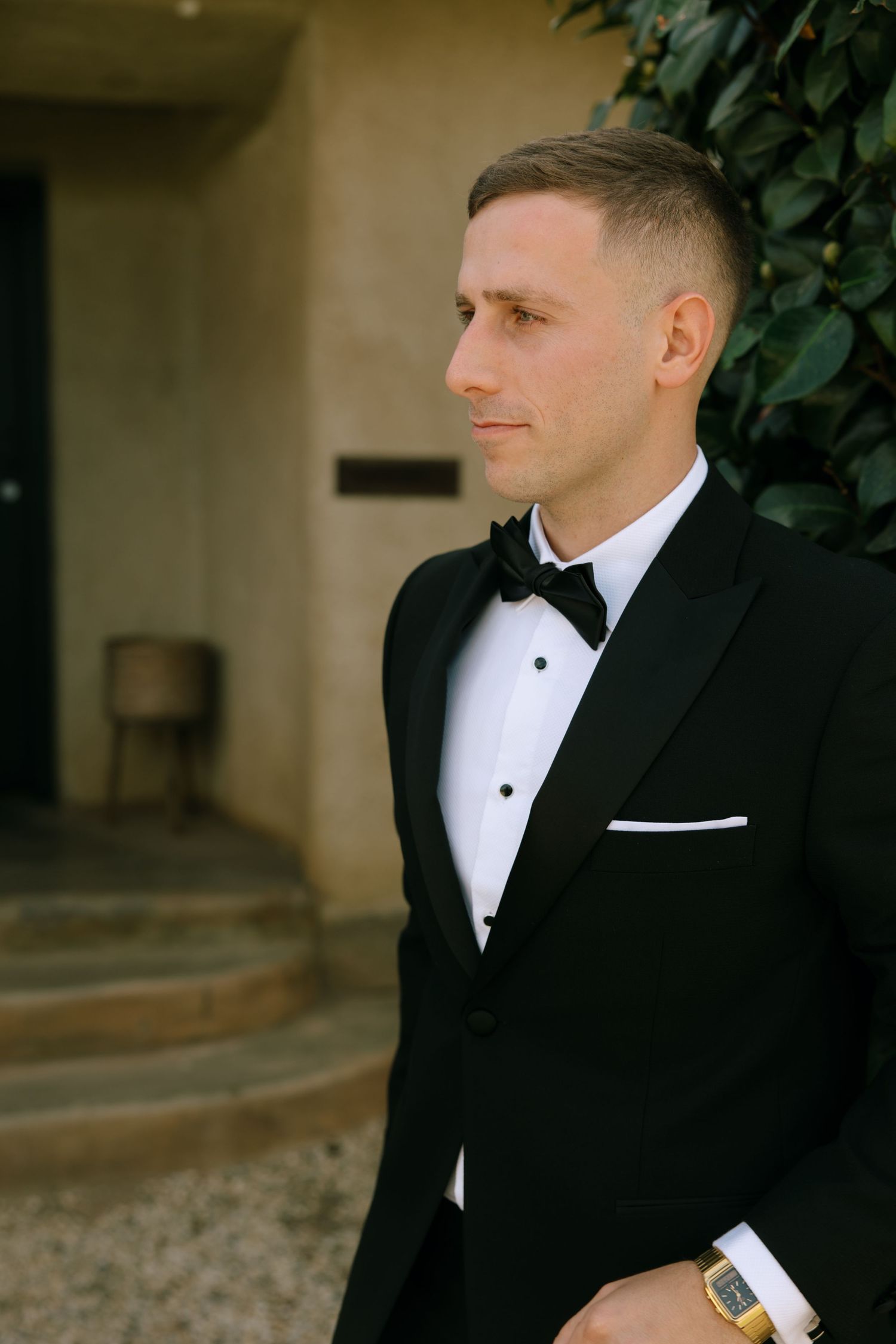Groom in classic black tuxedo and white bow tie poses thoughtfully against a rustic backdrop.