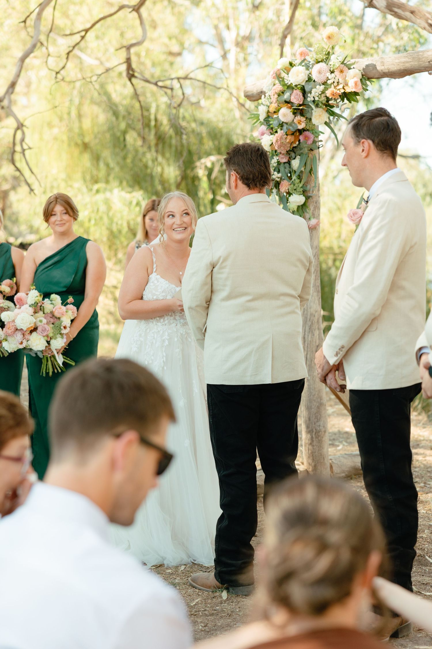 Intimate wedding ceremony in a wooded outdoor setting with bridesmaids in emerald green dresses and groomsmen in cream suits.