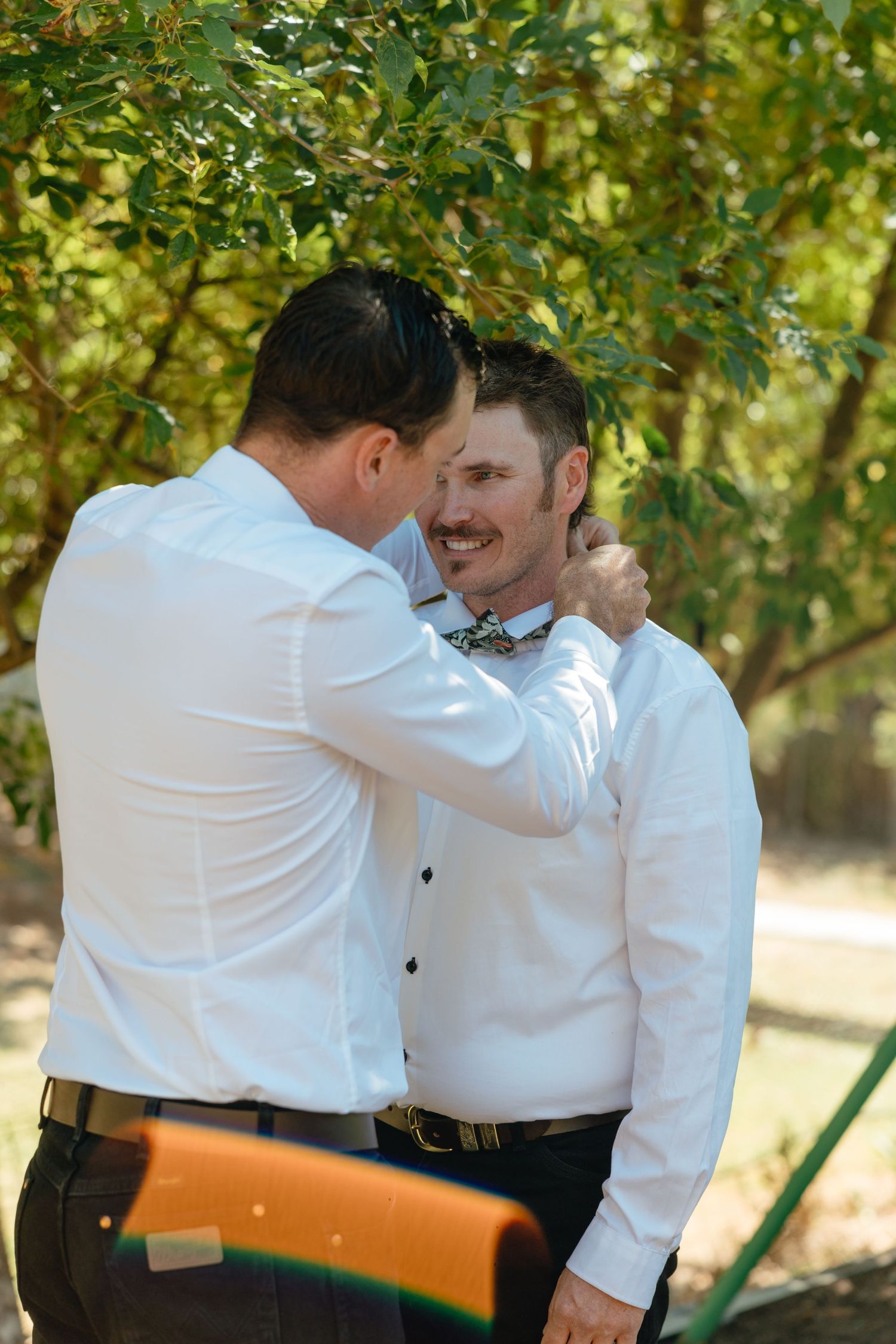 Two people in white dress shirts and bow ties share a tender outdoor moment under green foliage.