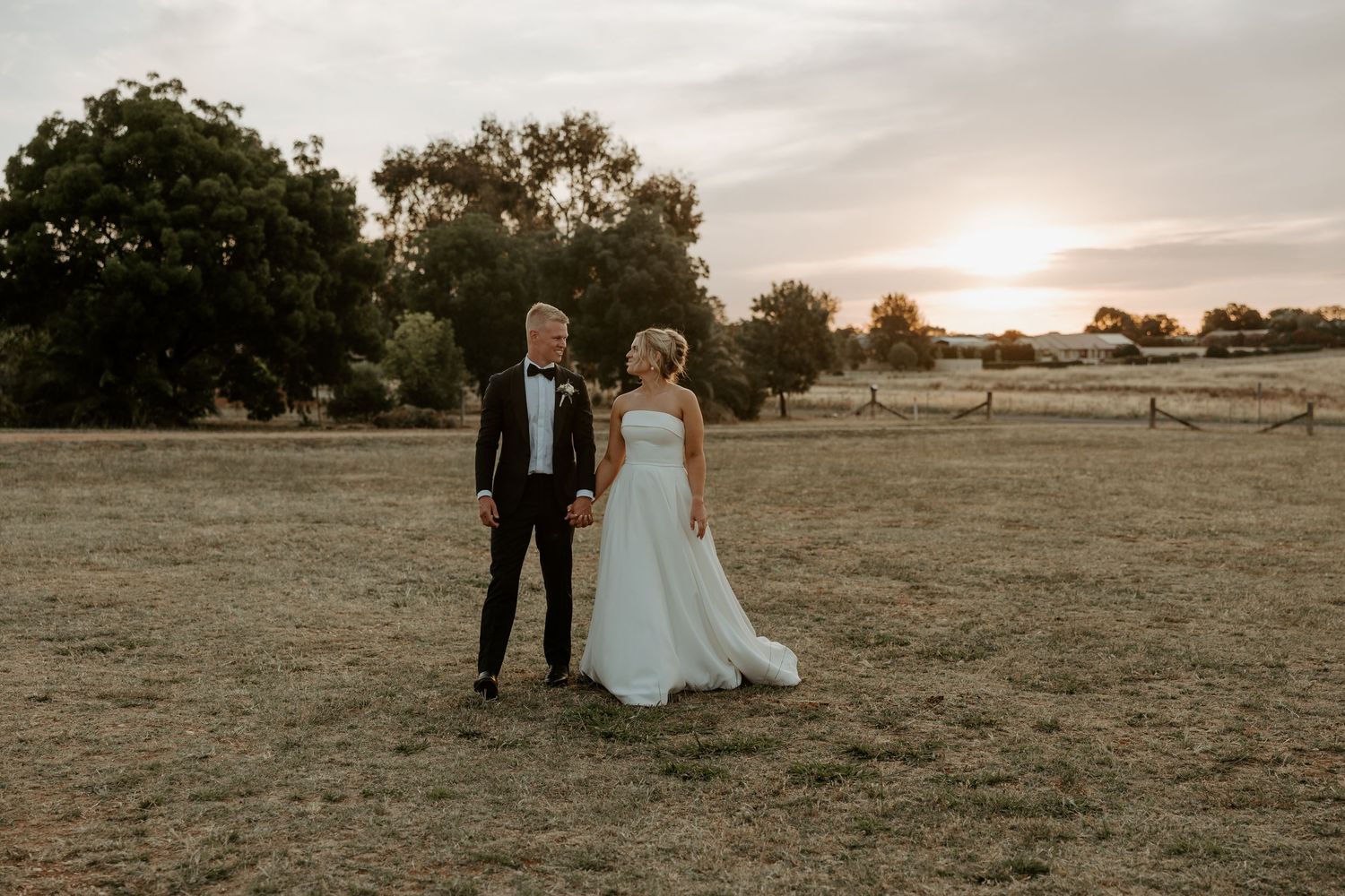 A couple stands together in a field at sunset with trees silhouetted in the background.