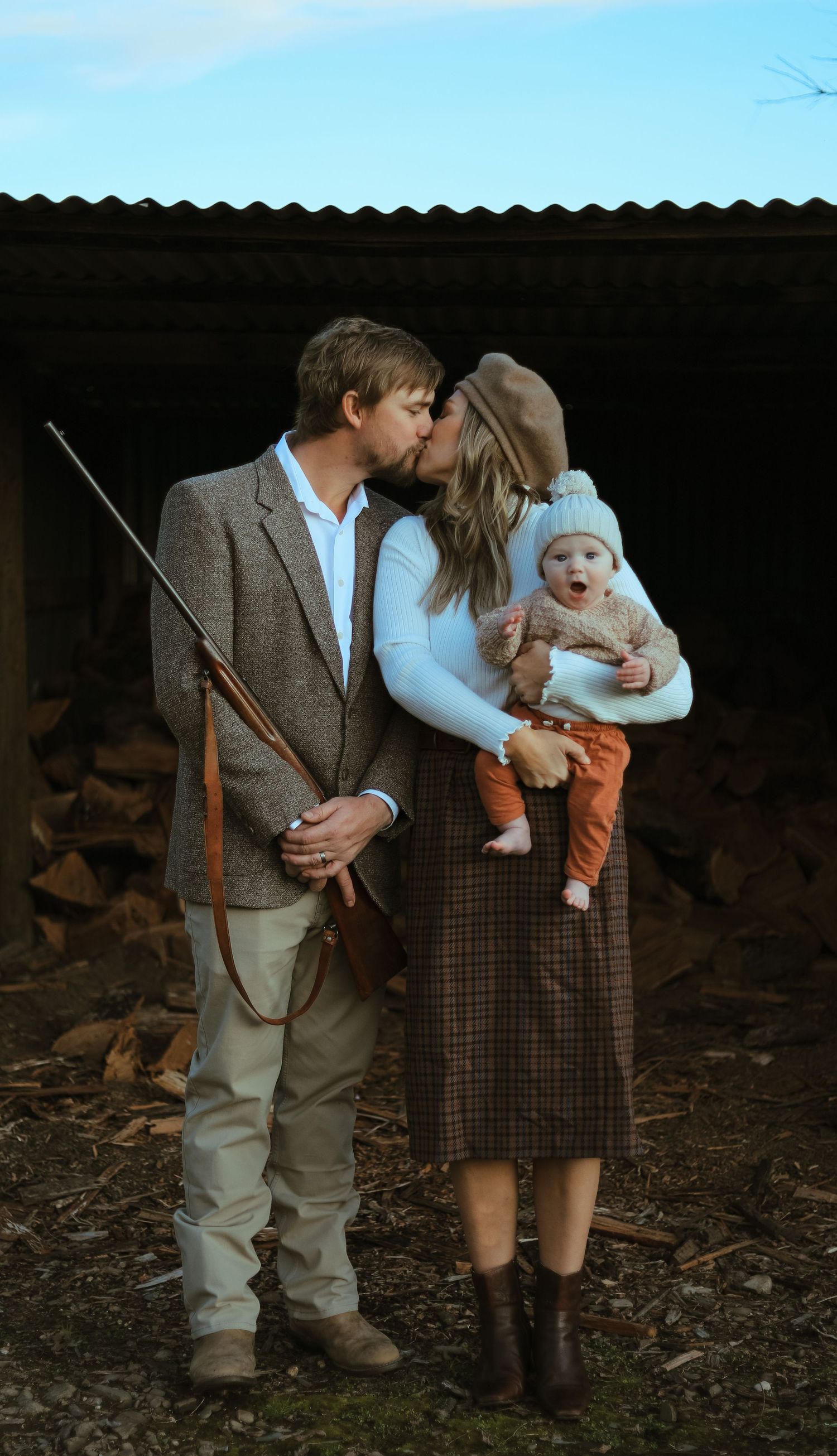 A couple kissing outdoors, woman holding a smiling baby, man carrying a rifle, wearing autumn country-style clothing.