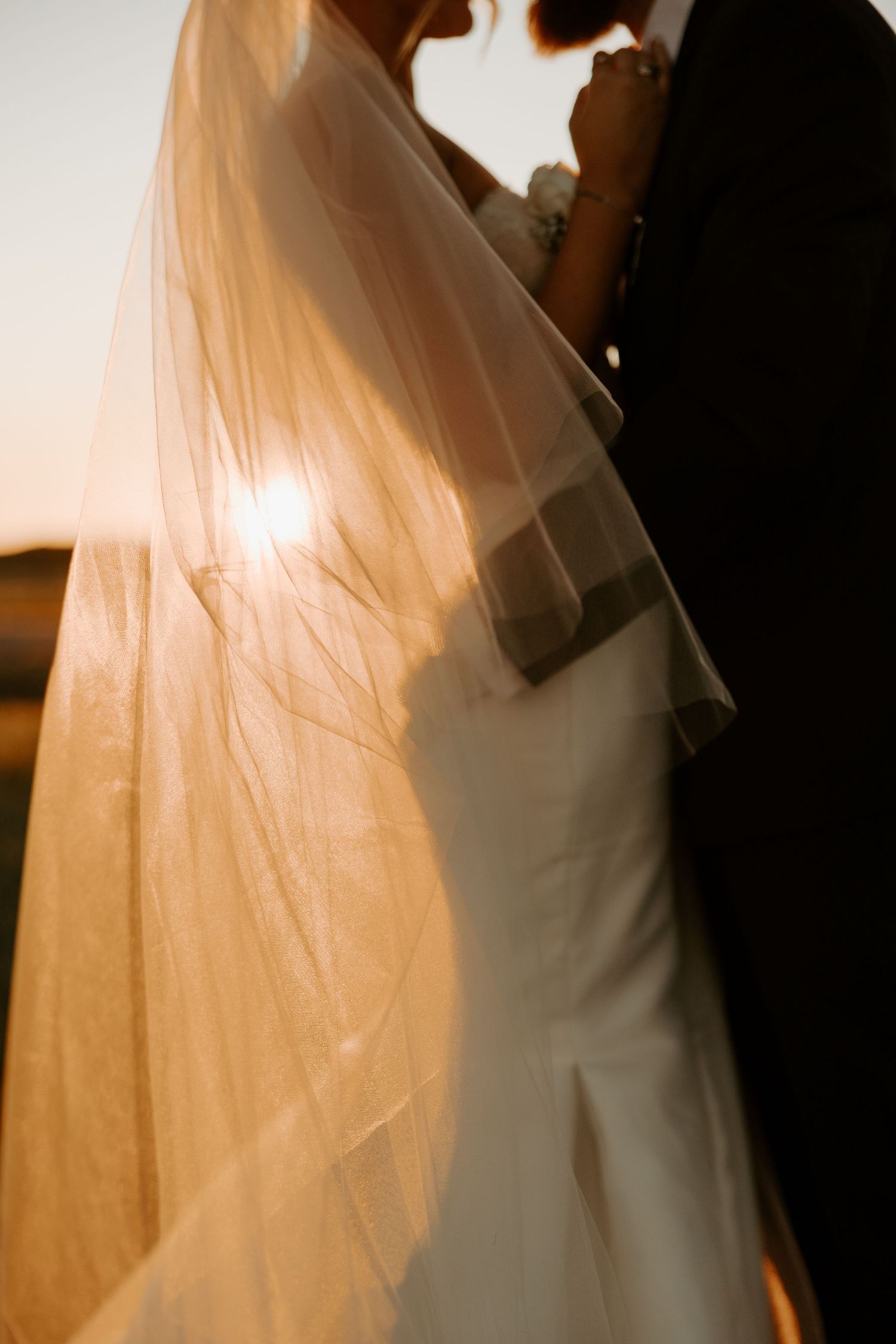Silhouette of wedding couple with flowing veil backlit by golden sunlight.