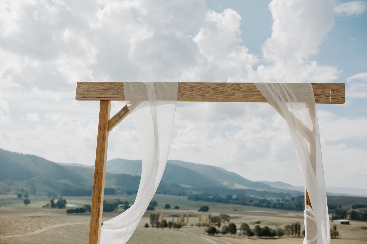 Wooden wedding arch with white fabric draping against mountain landscape.