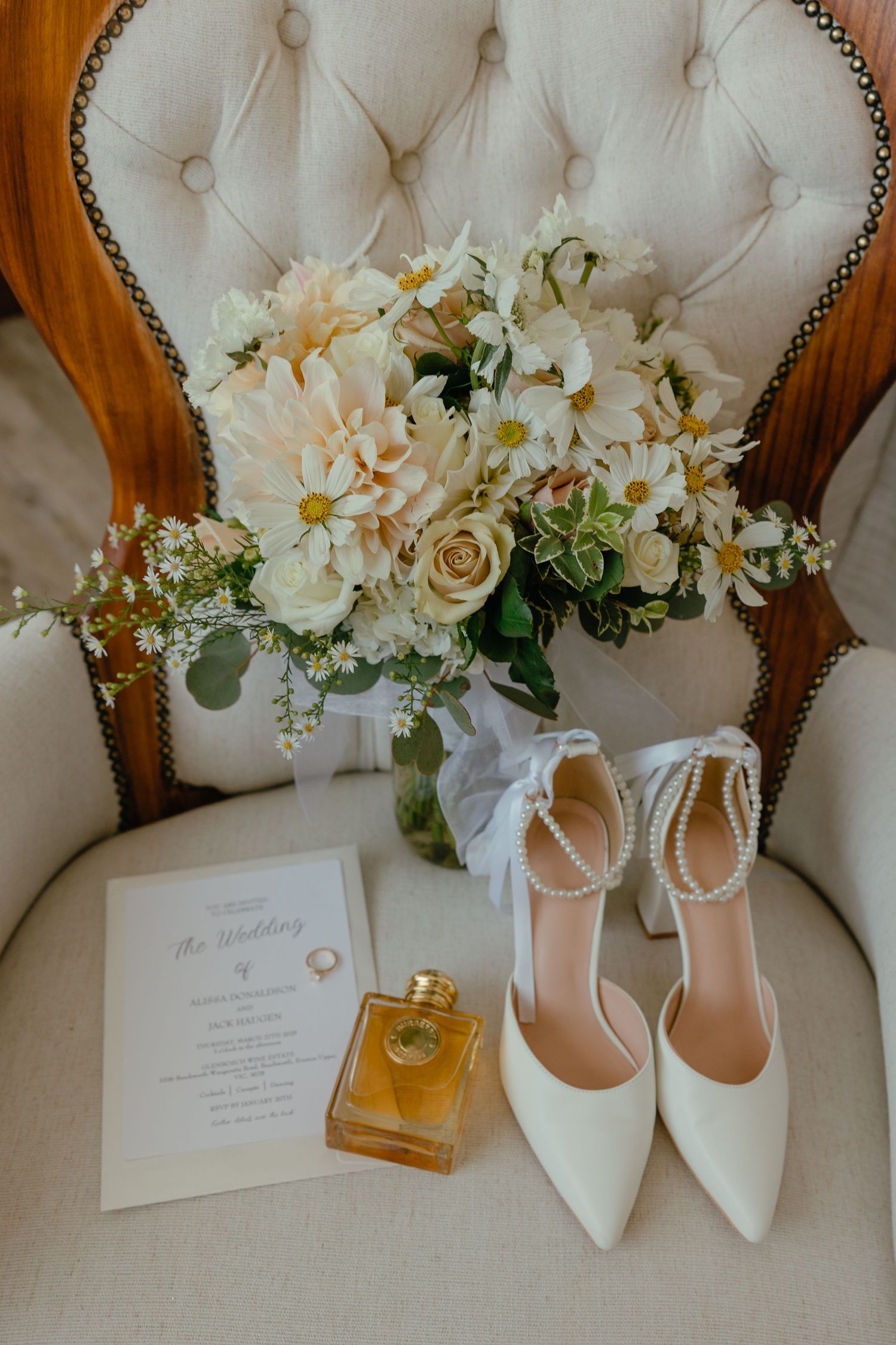 Wedding details flatlay with white heels, perfume bottle, invitation and cream floral arrangements on tufted white surface.