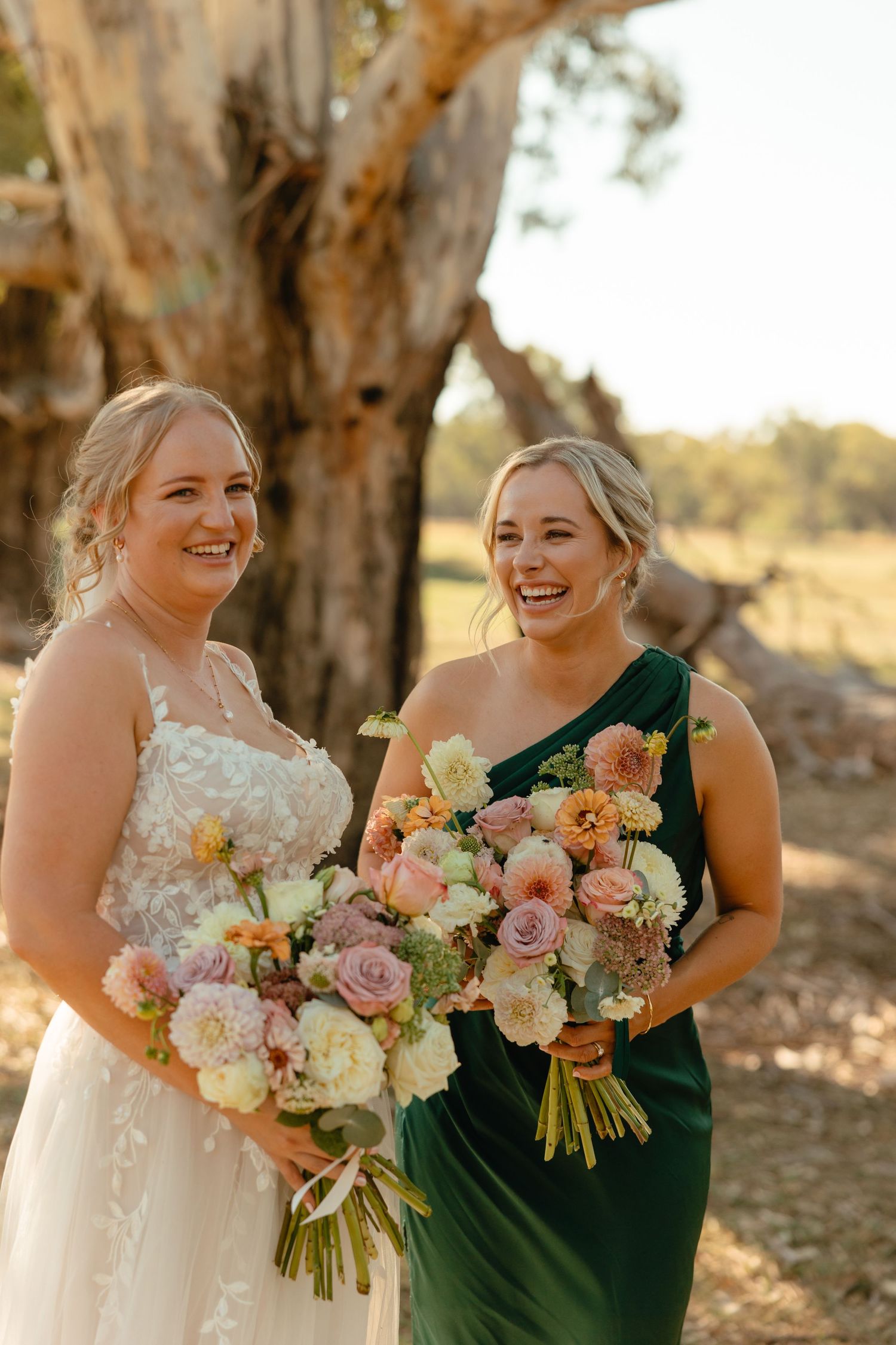 Wedding party members share candid laughs while holding beautiful bouquets of pink and cream roses against sunset backdrop.