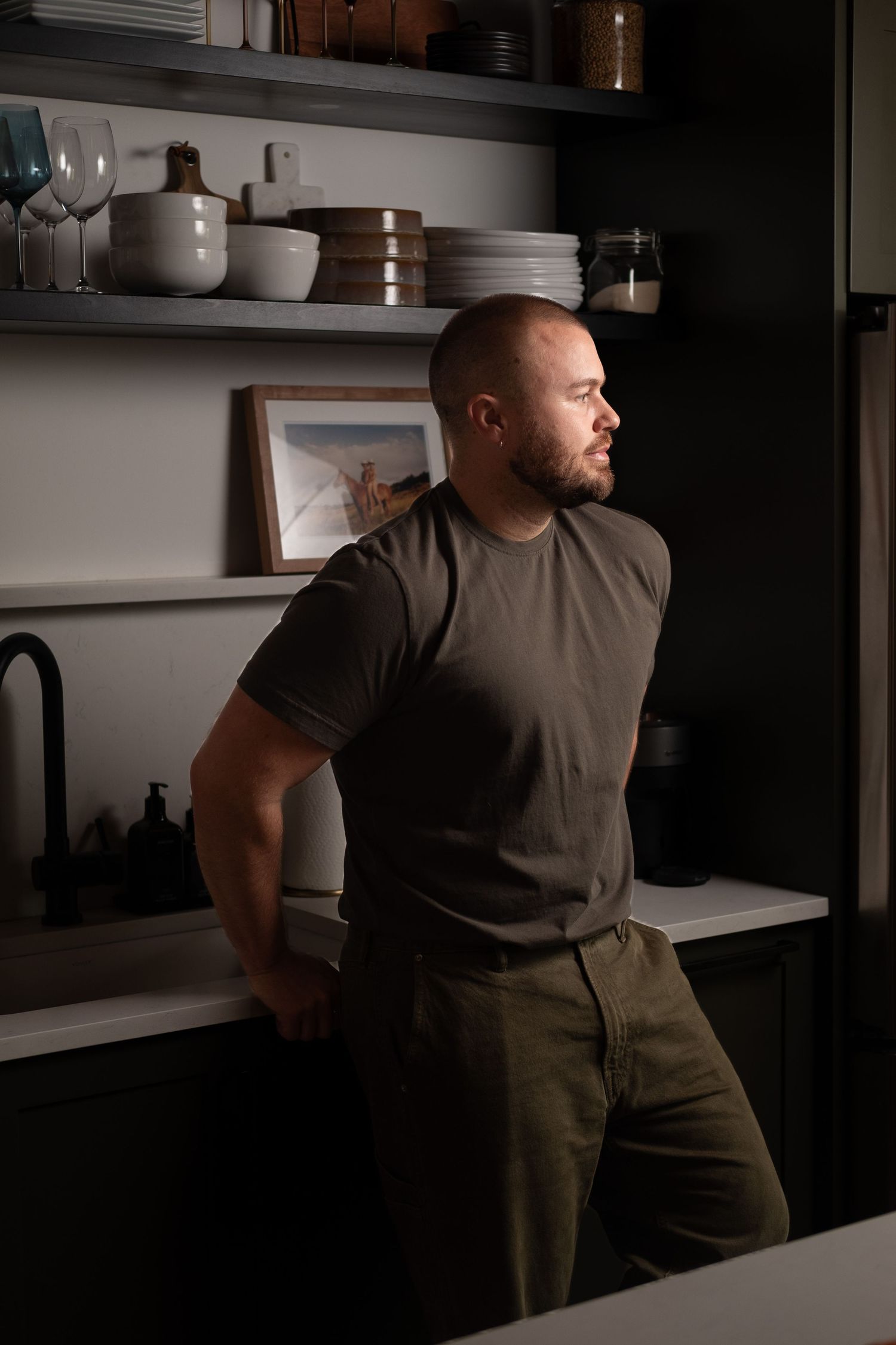 A man take a digital photographer in his kitchen. He is wearing green.