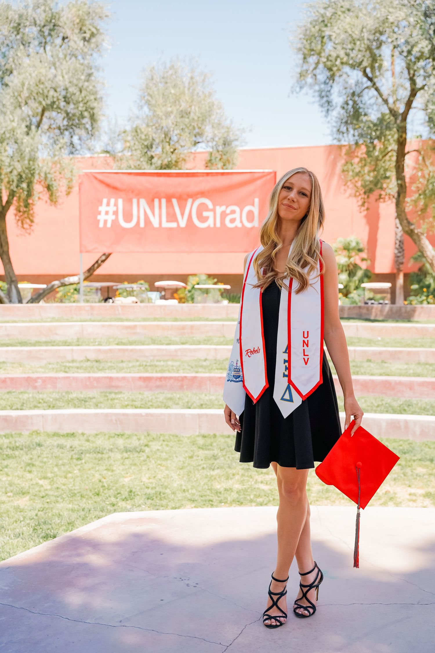 Graduate in black dress and graduation stoles poses by UNLV sign during graduation photos.